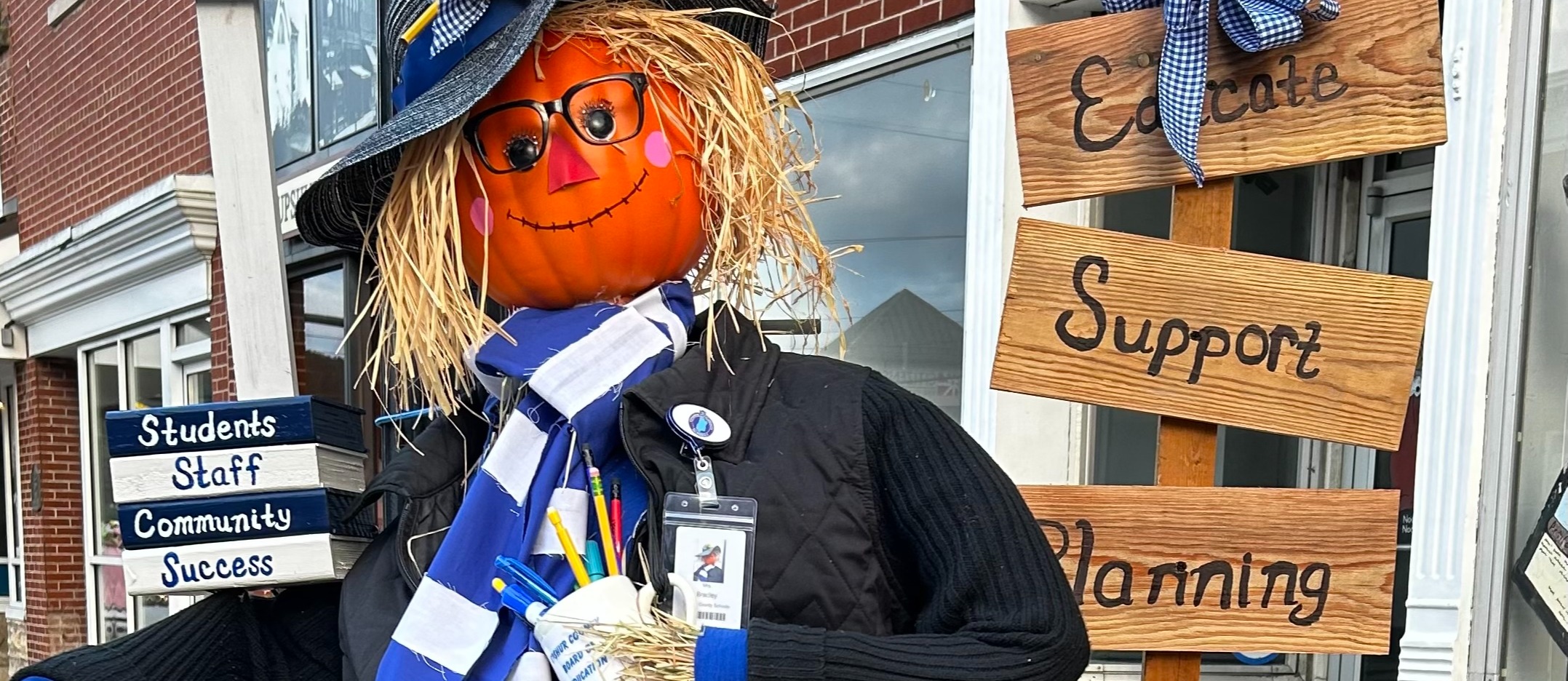 A fall-themed scarecrow display stands on a downtown sidewalk in front of a brick building. The scarecrow has a pumpkin head, straw hair, glasses, a black hat decorated with a sunflower, a blue-and-white scarf, and a school ID badge. It holds a sign reading “Students, Staff, Community, Success.” Beside the scarecrow, wooden signs list “Educate,” “Support,” “Planning,” “Partnerships,” and “Better Tomorrows.” A flower pot filled with colorful autumn flowers displays an “I ❤️ WV Public Schools” logo. The display is decorated with sunflowers, straw, and fall foliage.