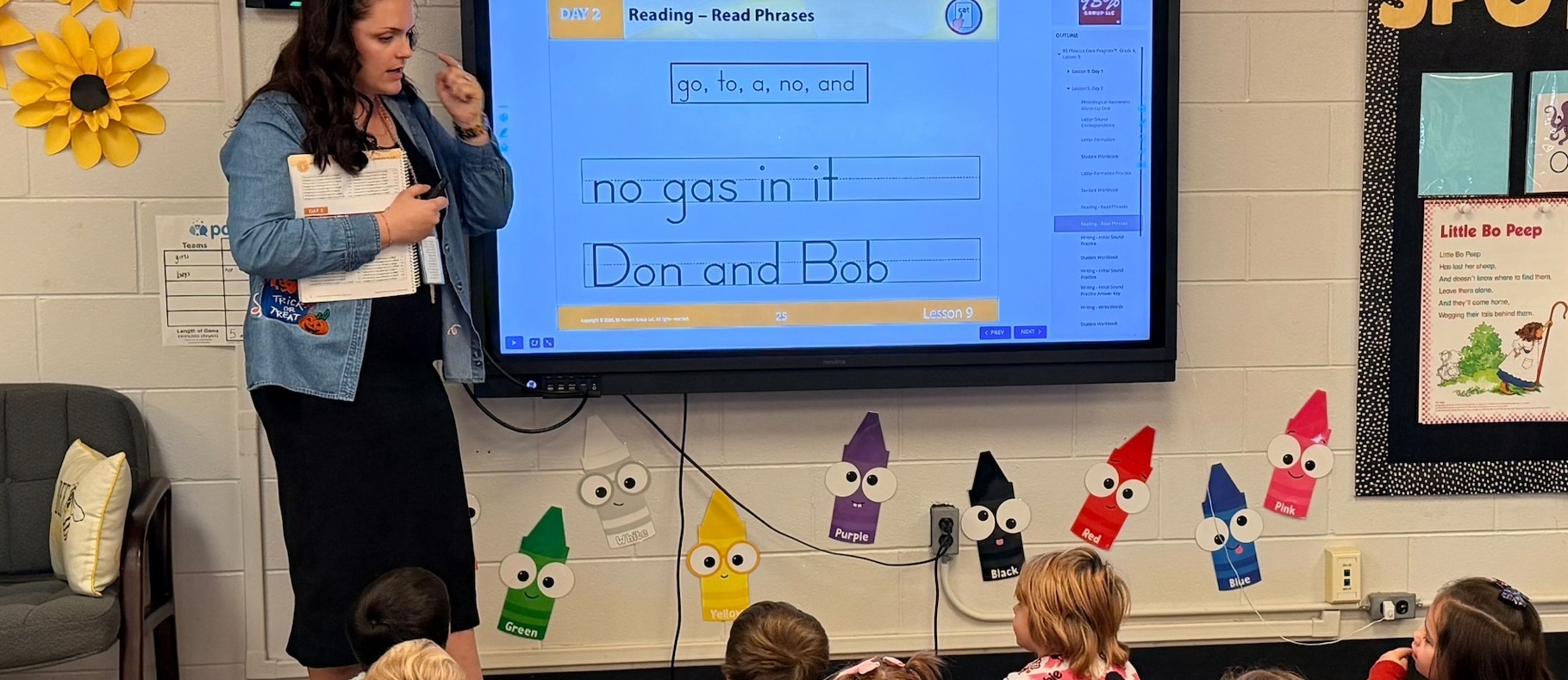 A teacher stands beside an interactive classroom display while leading a small-group reading lesson with elementary students seated on a blue carpet. The screen shows a phonics and reading exercise titled “Reading – Read Phrases,” with phrases including “no gas in it” and “Don and Bob.” Students face the display and teacher, participating in the lesson. The classroom features bright yellow sunflower decorations, an alphabet display hanging across the wall, colorful crayon-shaped cutouts labeled by color, and a literacy bulletin board displaying the nursery rhyme “Little Bo Peep.” The room is well-organized and designed to support early literacy instruction through direct teaching and student engagement.