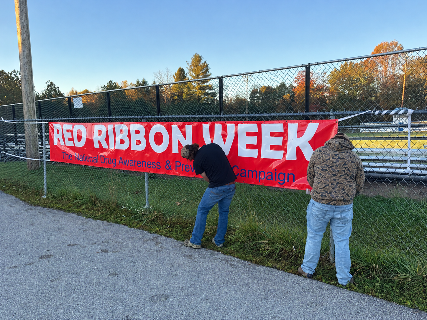 Two high school students attach a large red banner reading “Red Ribbon Week: The National Drug Awareness &amp; Prevention Campaign” to a chain-link fence beside an athletic field. The photo was taken outdoors on a clear autumn morning, with bleachers, trees displaying fall colors, and a blue sky visible in the background. The students are securing the banner as part of Red Ribbon Week awareness activities on campus.