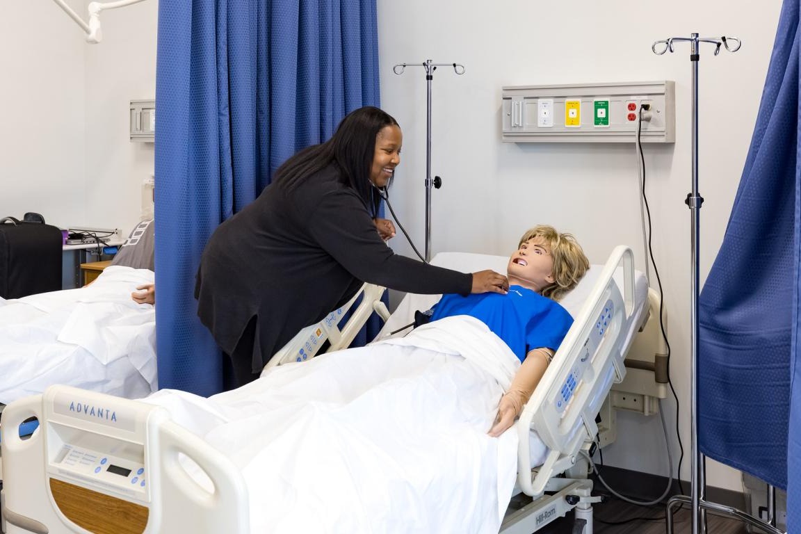 A woman in a black coat helps a patient in a blue shirt in a hospital bed.