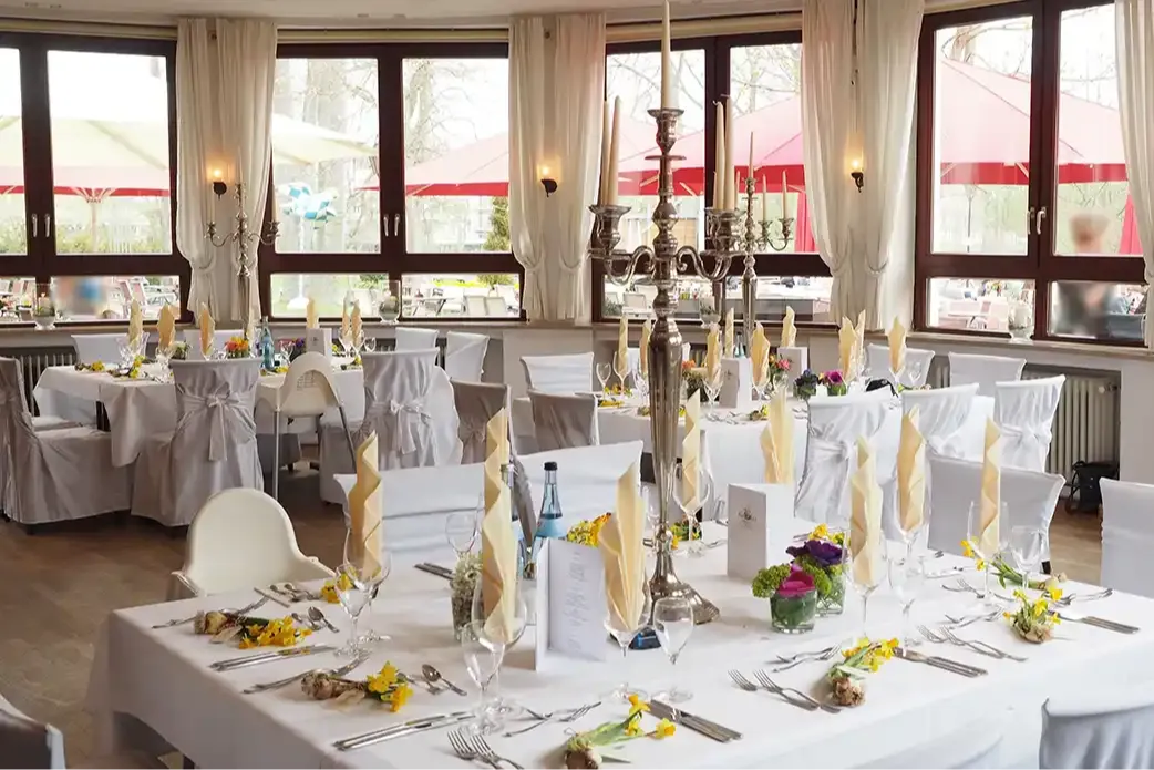 Large room with large windows and white curtains. A long table set with white cloths, flowers, and silverware.