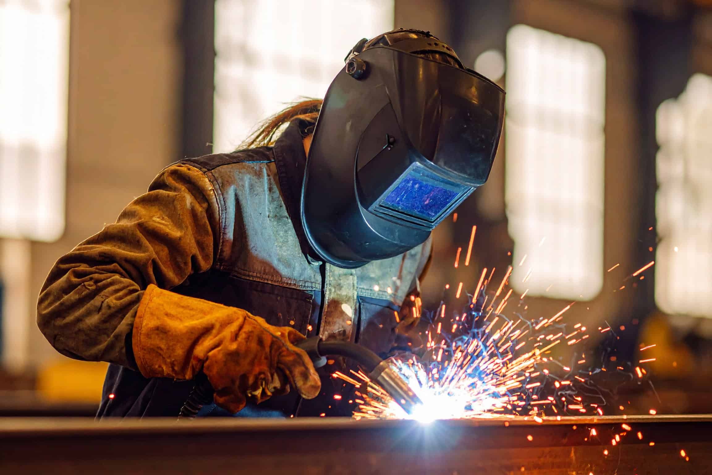 A person wearing a welding helmet and gloves welds metal in a factory with large windows.