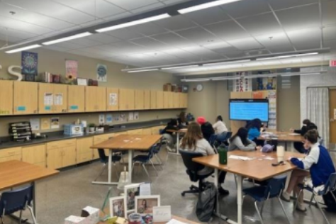 A classroom with desks and chairs, a large screen on the wall, and overhead lighting. Students sit at desks.