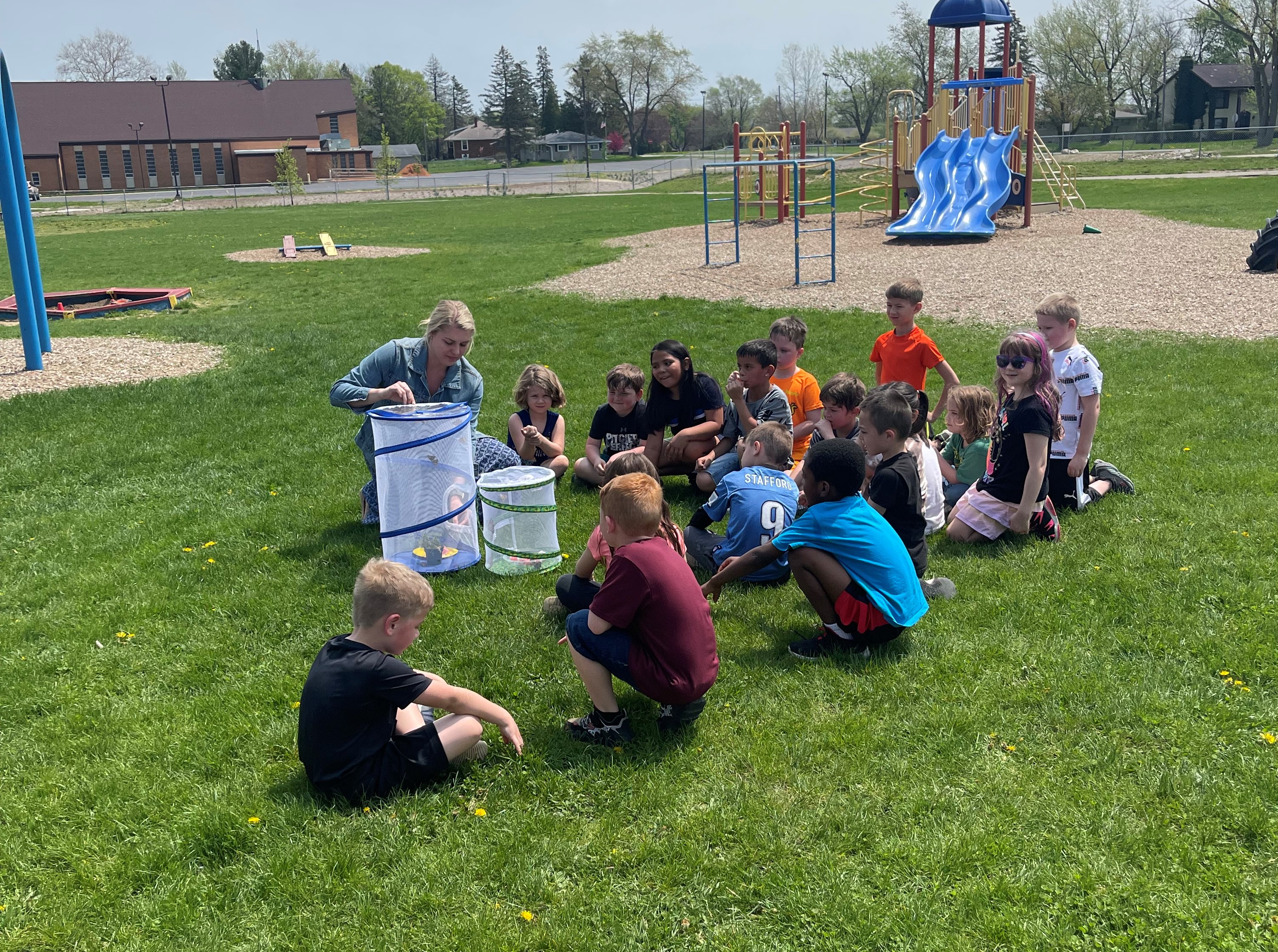 A group of about  15 students sit outside on the lawn while a teacher shows butterflies in butterfly houses.  There is a playground in the background.