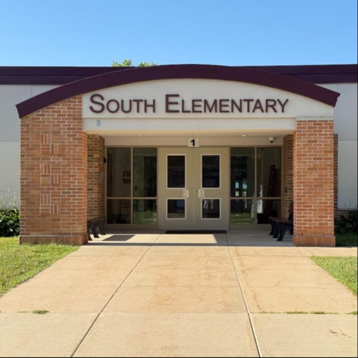 Entrance to South Elementary. A concrete walkway leads to double doors. Brick pillars support a sign.
