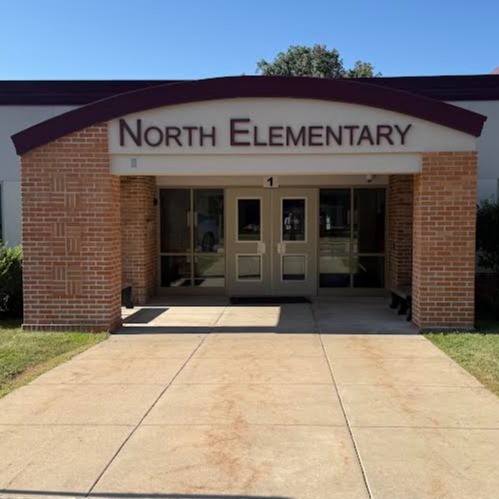 A building with a sign reading "North Elementary" in front of it. It has brick columns, double doors, and is surrounded by grass and bushes.