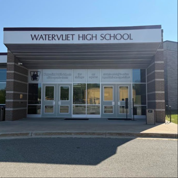 Entrance to Watervliet High School, featuring a sign, glass doors, and a paved area. Behind the school is a grassy lawn.