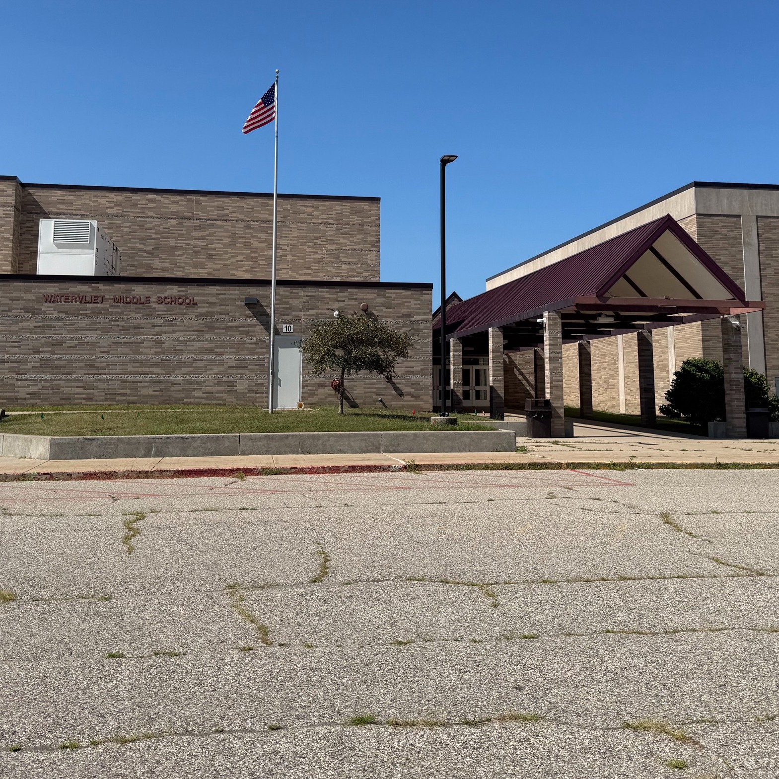 A school building with a flag in the foreground. Behind it, an entrance with a canopy. Trees and plants are nearby.