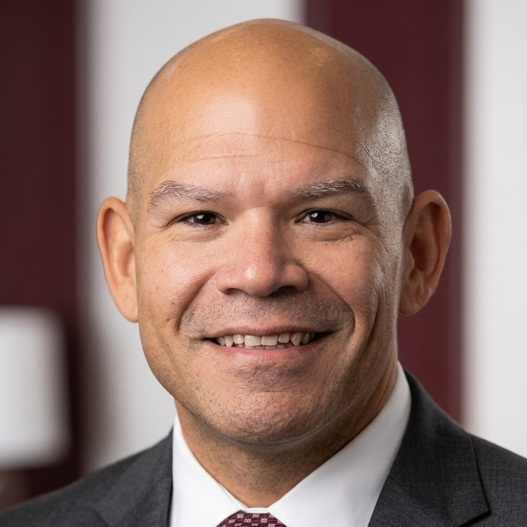 A man with a bald head, wearing a gray suit and a maroon tie, smiles at the camera.