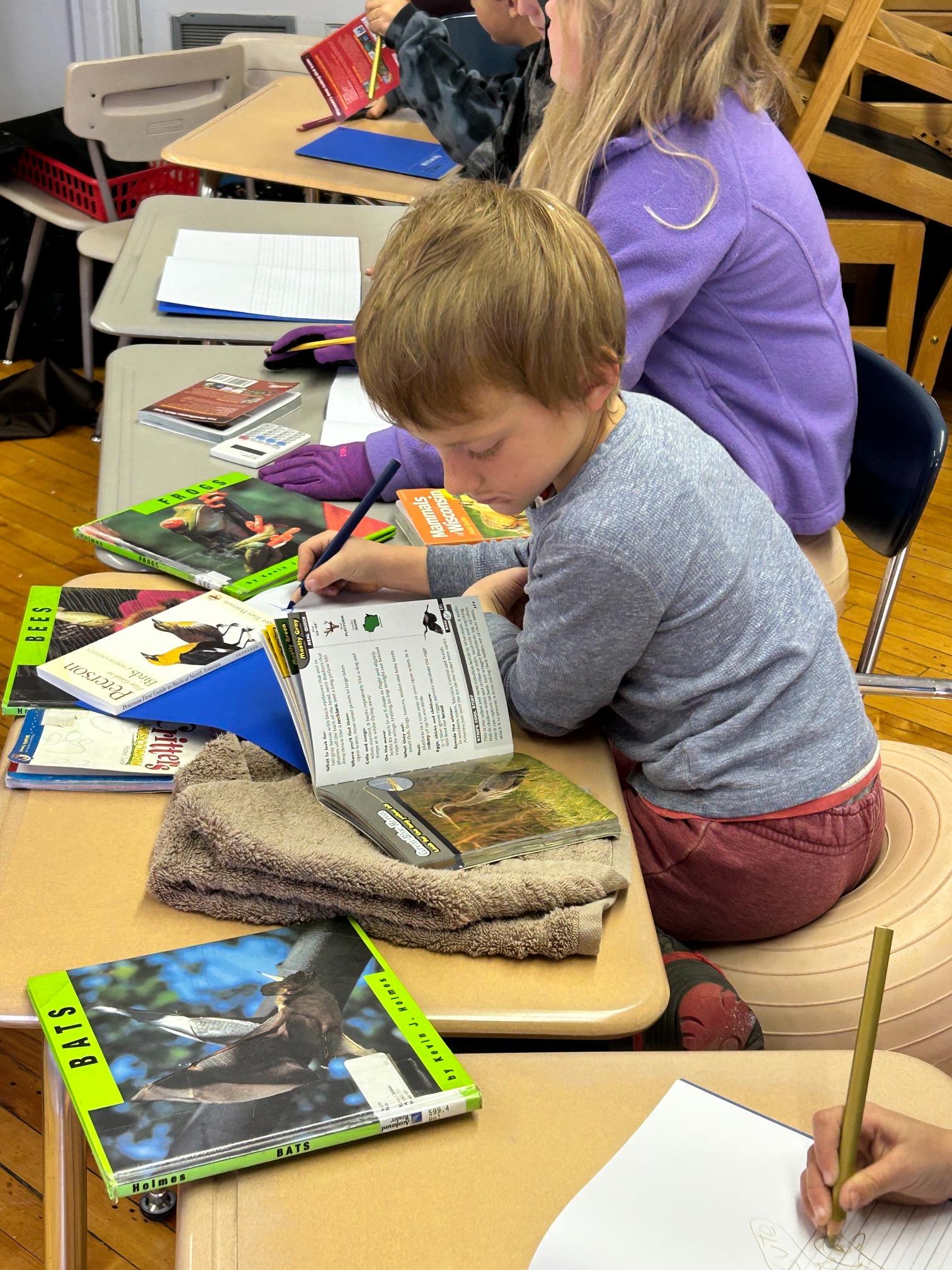 A child sits at a desk with an adult, both engaged in writing. Books, papers, and other items are on the desk.