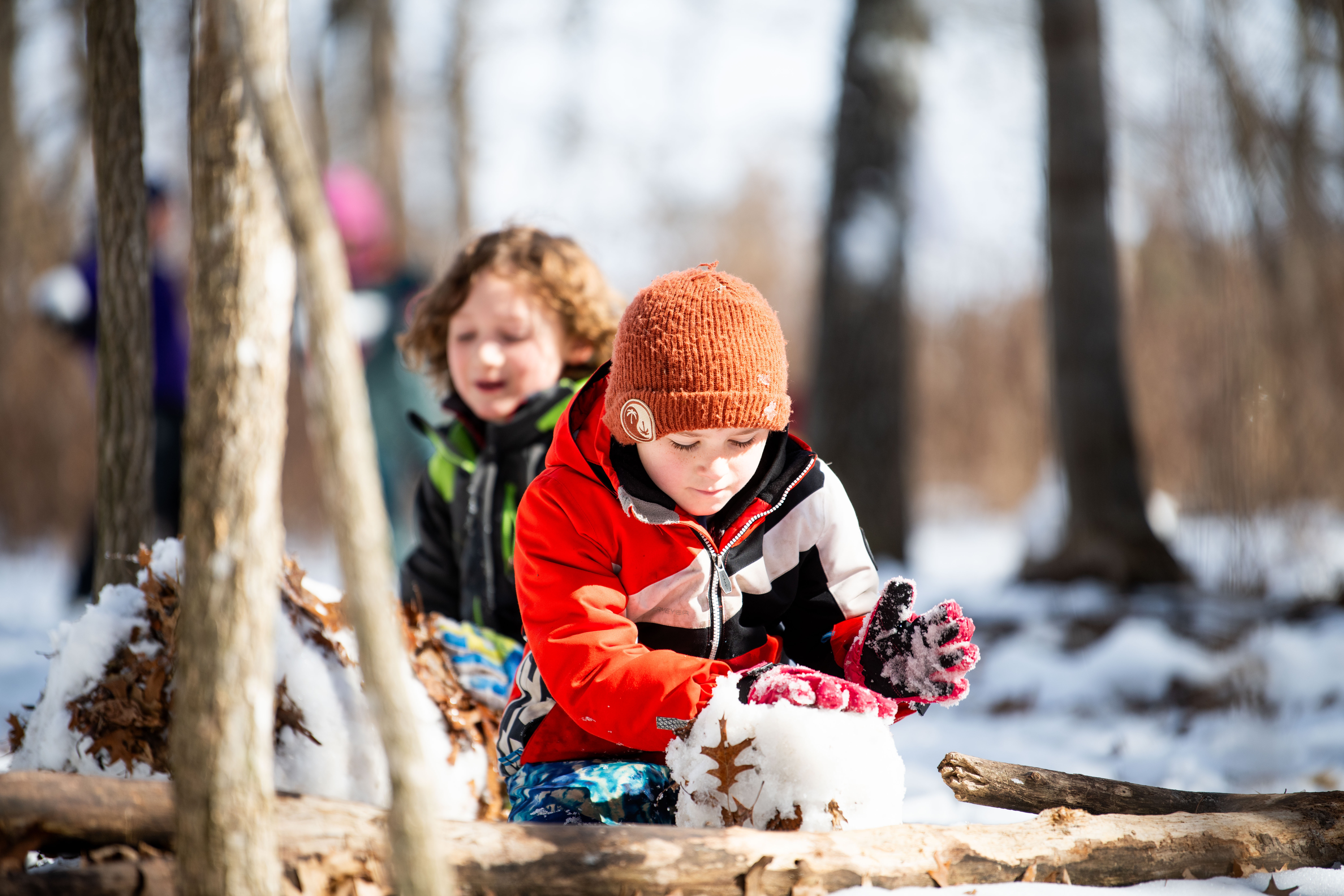 Two children playing in the snow. One is wearing a red hat and holding a snowball.