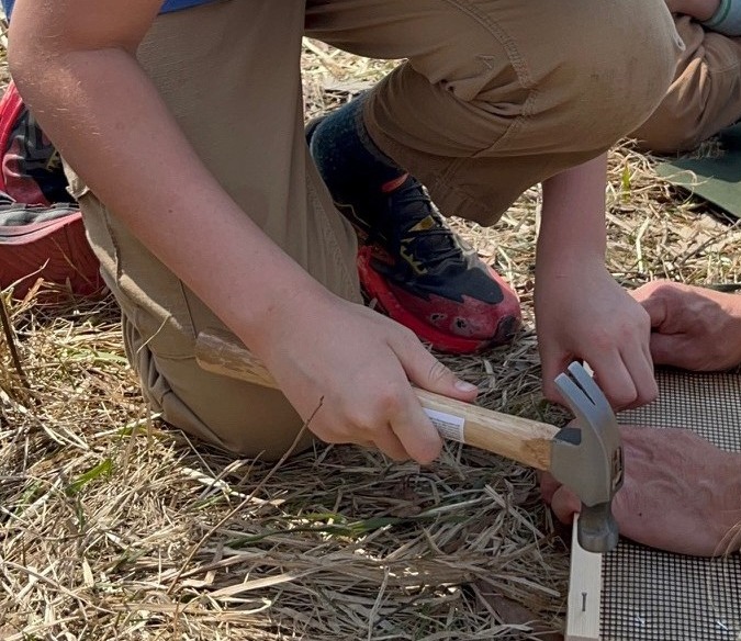 People outdoors, one holding a hammer and working on a wooden plank, another person holding the plank.