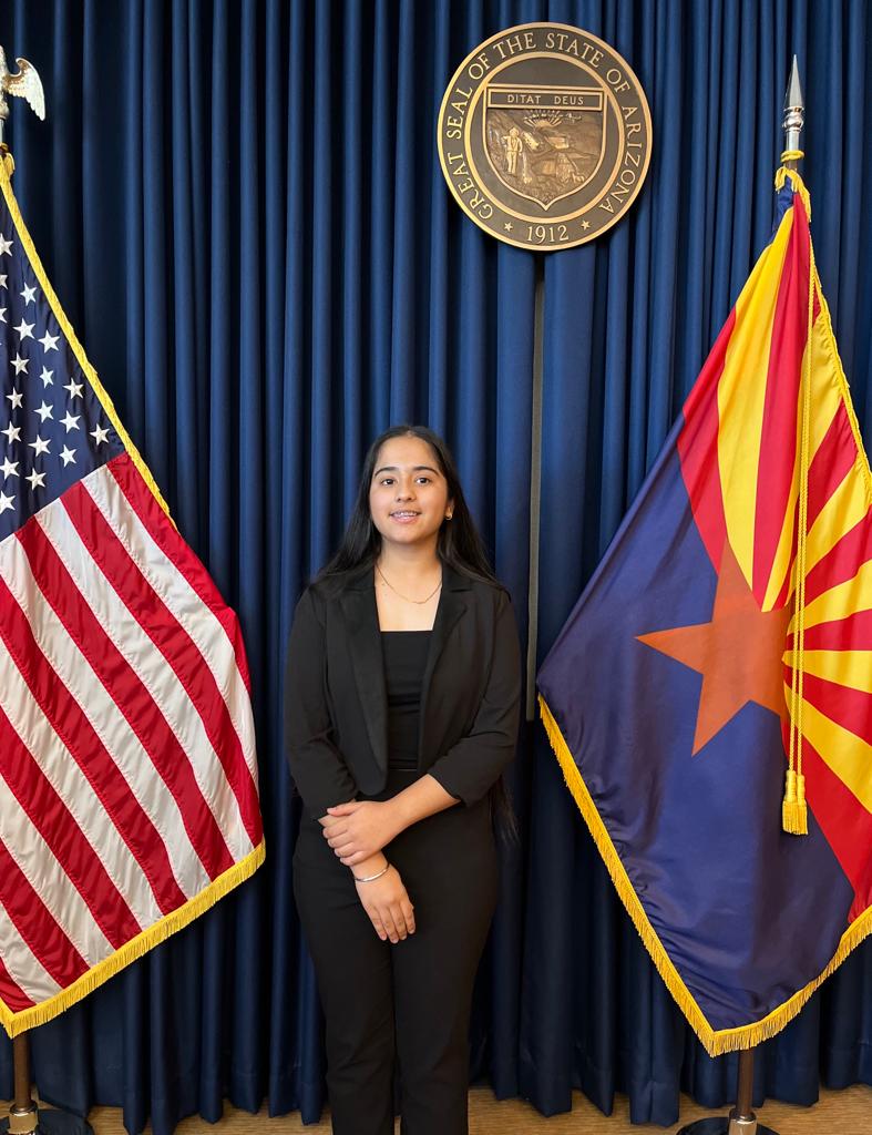 Ms. Kaur poses against a blue background flanked by the United States and Arizona flags