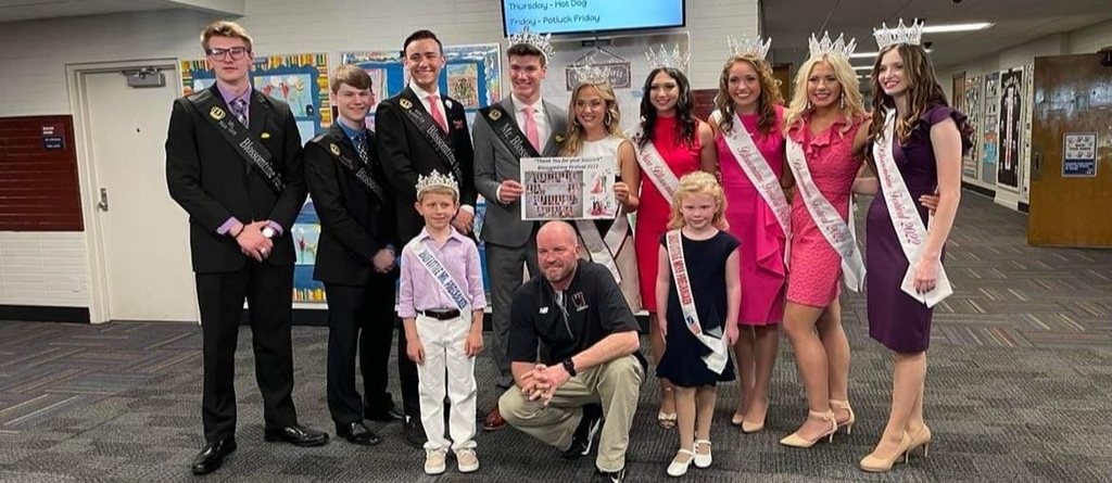 Principal Bill Tiefenbach crouches down to the level of the the little Blossomtime Prince and Princess while the other Blossomtime Royalty stand behind them, all dressed nicely and wearing their crowns