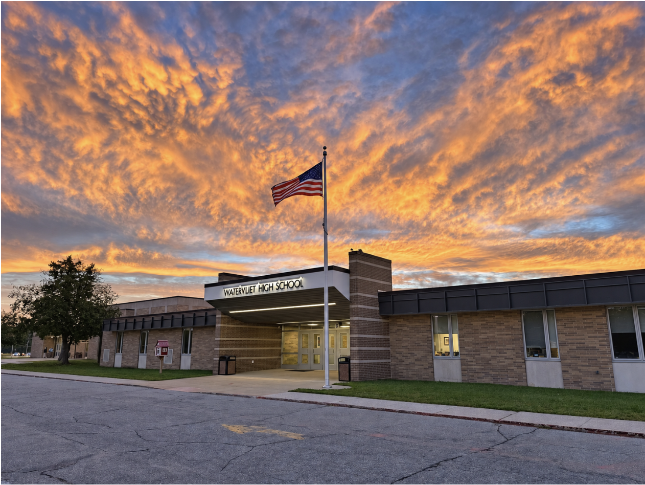 Sunset behind school building with flag on pole; building has brick walls, glass windows, and a large entrance.