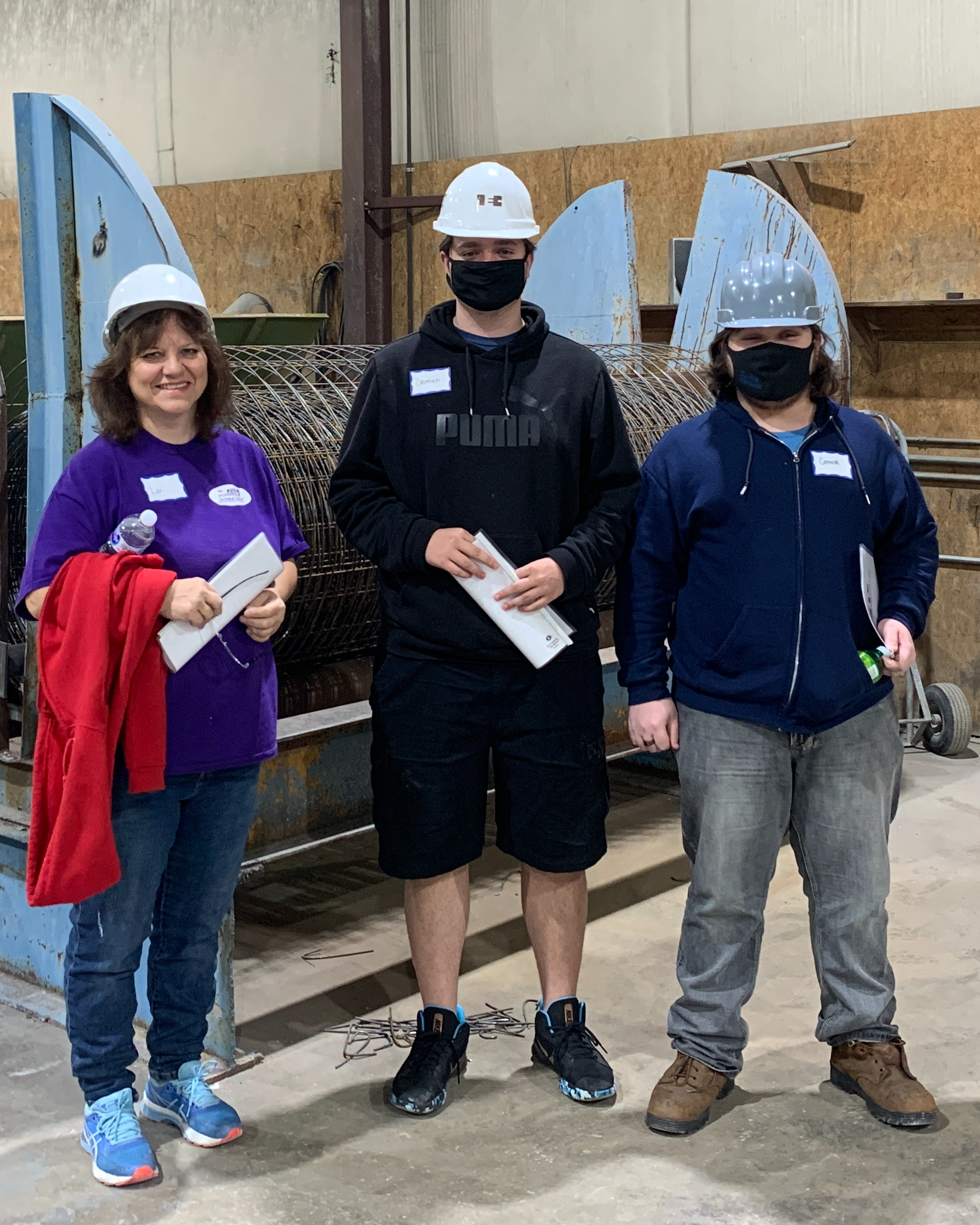 Three people in hard hats and masks stand in front of large blue machinery. One holds a red jacket.