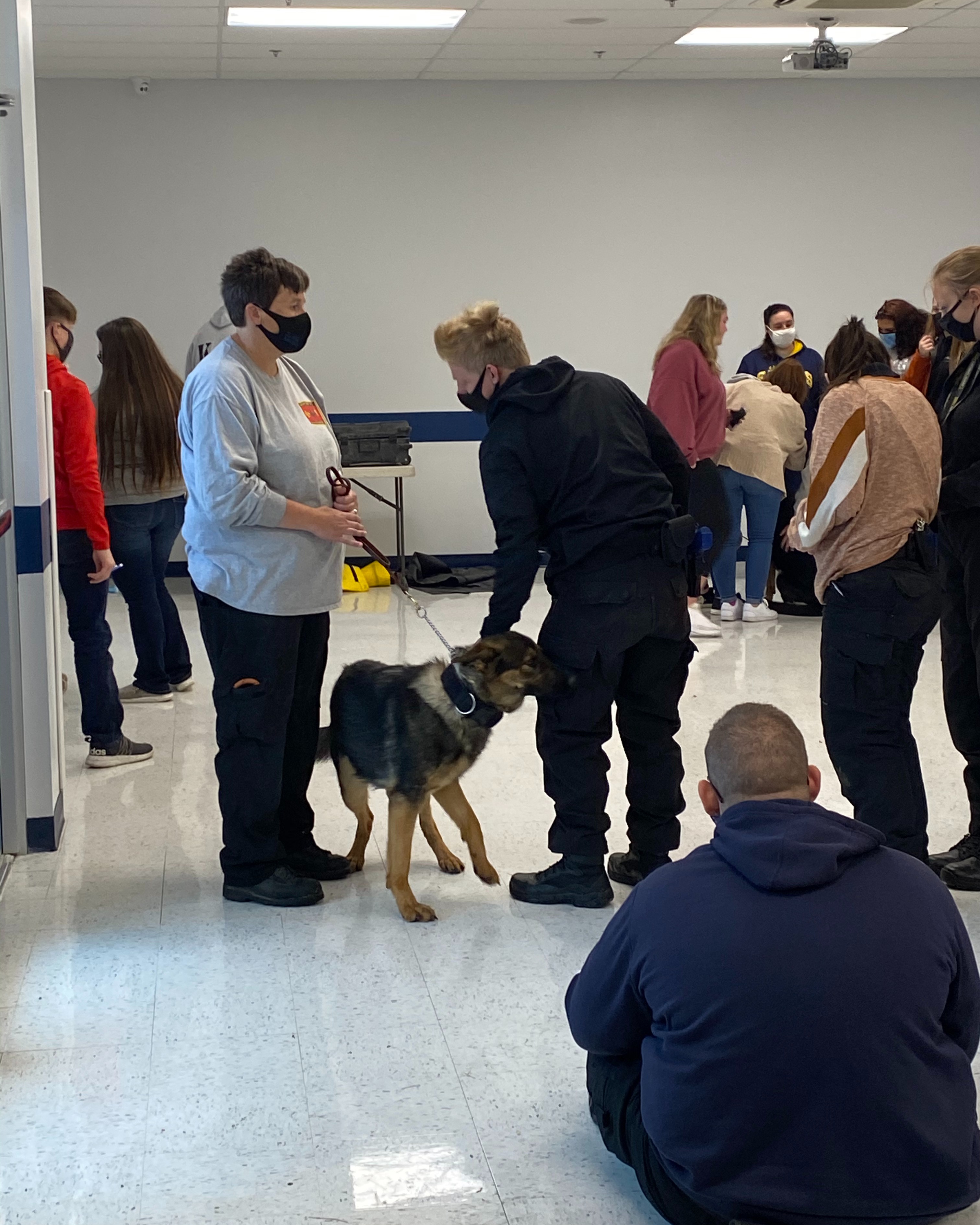 People in a room with white walls and floor. A dog stands near two adults. One holds a leash.