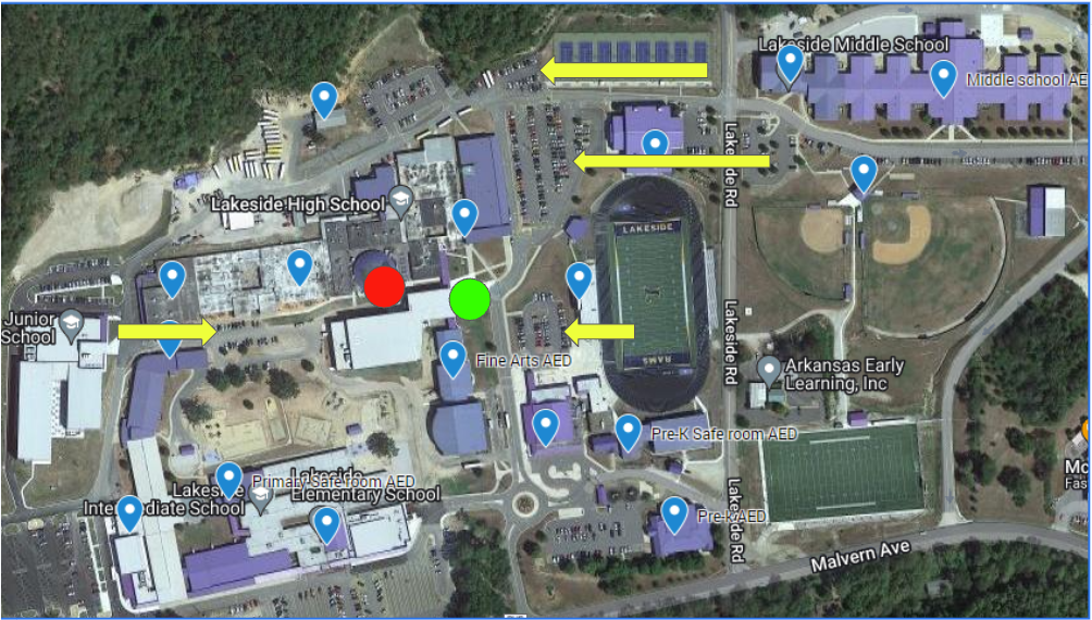 Annotated aerial map of the Lakeside campus. School buildings are shaded purple, and blue map pins mark AED locations across the campus. Several yellow arrows show directions along campus roads and parking areas. A red dot marks a location near the central buildings west of the stadium, and a green dot marks another nearby location closer to the Fine Arts AED pin. The football stadium is in the center-right of the image, with baseball fields above and to the right and additional school buildings spread across the campus.