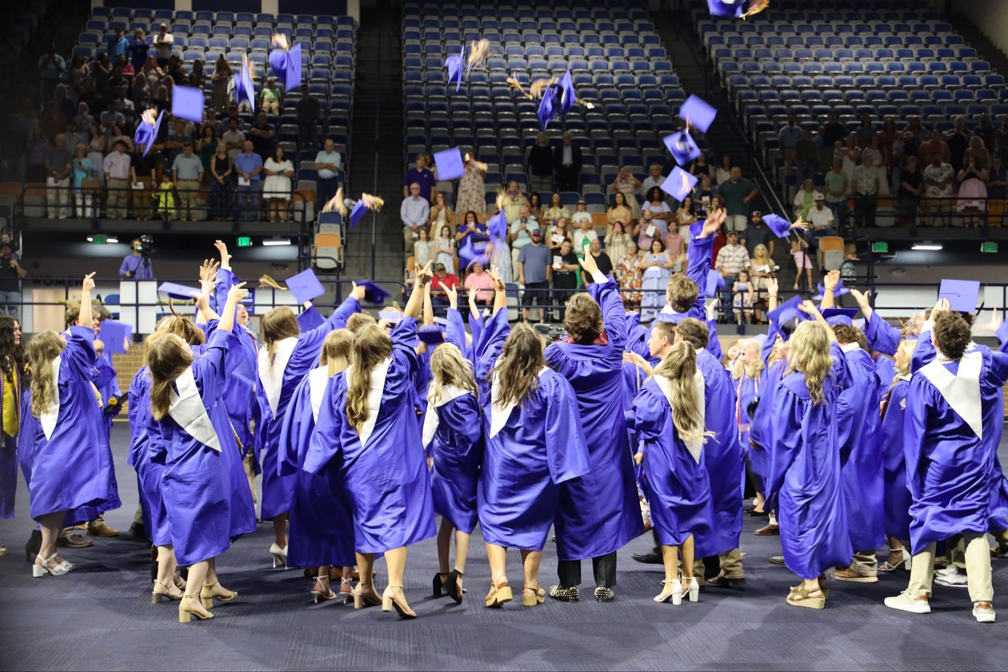 Graduates in blue gowns and caps throwing them up on a stage in a stadium.