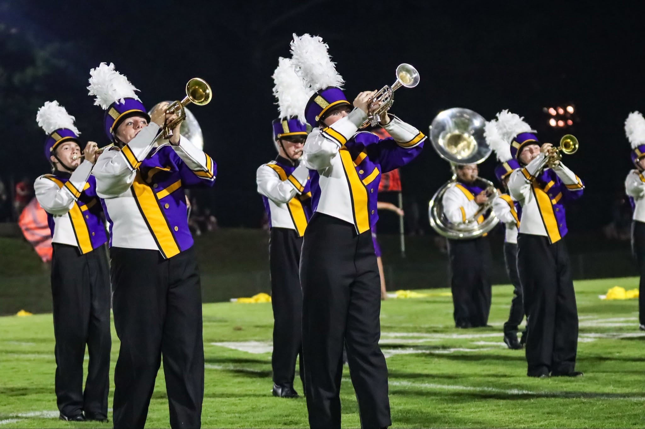 A marching band performs on a football field at night. Members wear purple and white uniforms with white hats.