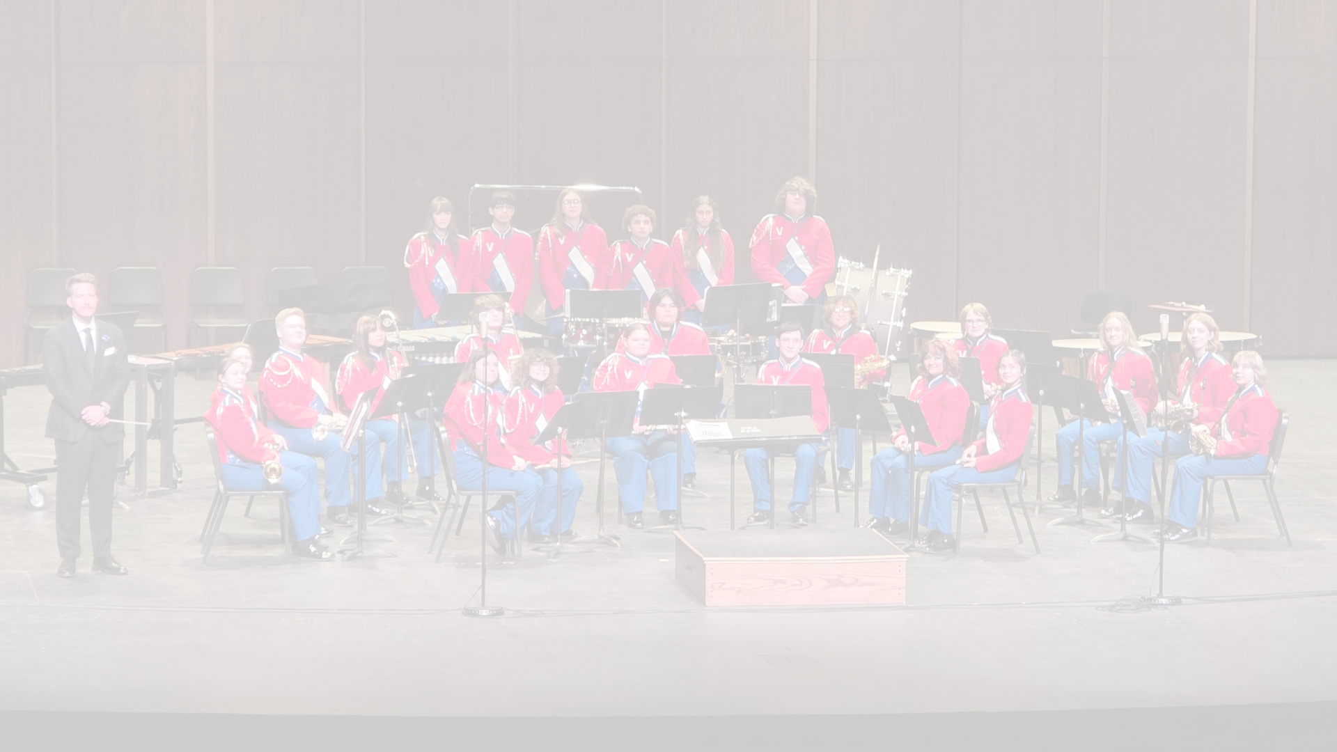 A group of people in red uniforms playing instruments, seated on chairs, with a conductor standing on a stage.