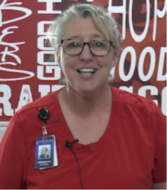 A woman in red scrubs smiles and wears glasses and a badge in front of a banner.