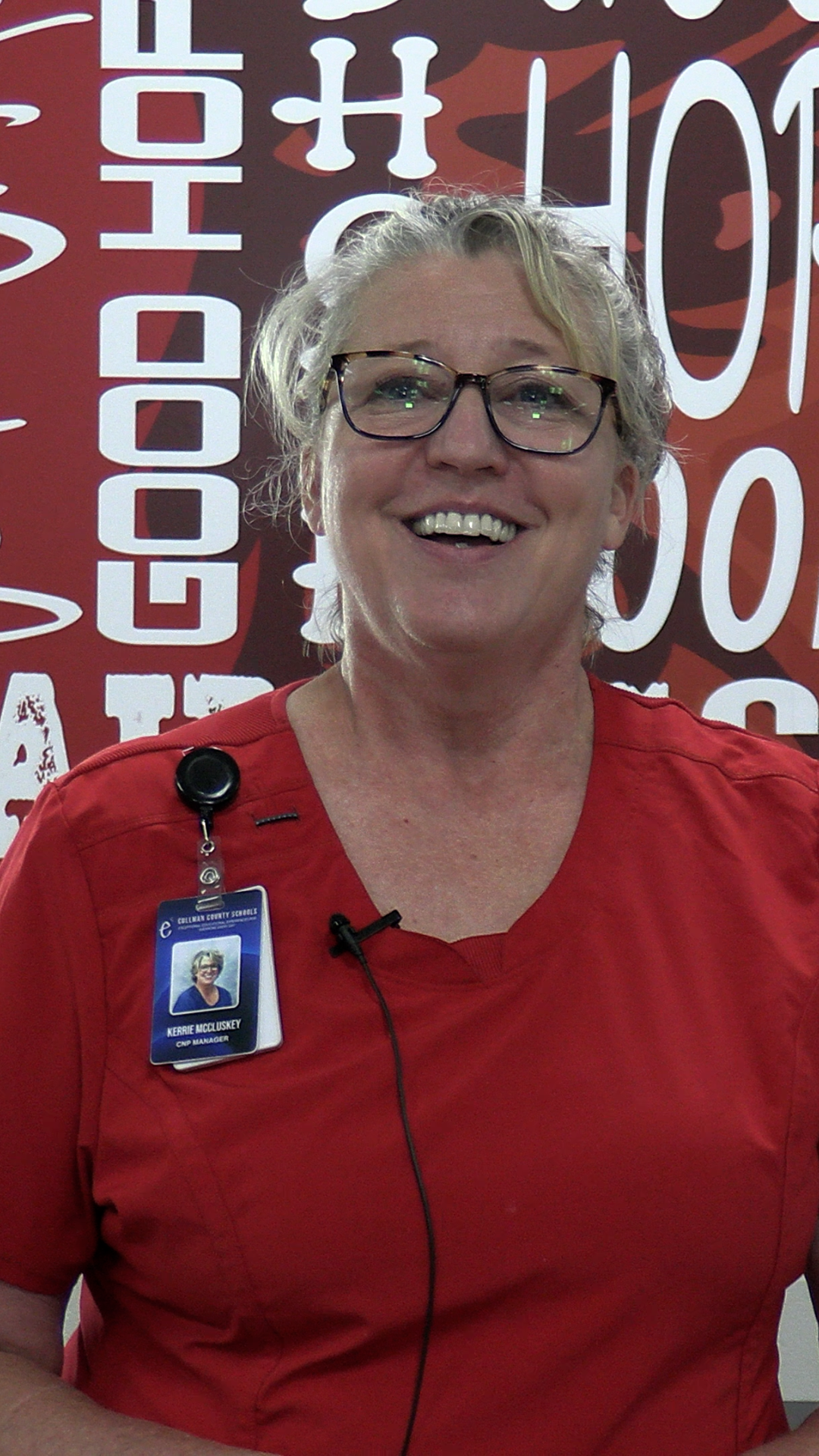 A woman in a red shirt with a name tag smiles against a wall with "Good Hope" and other text.