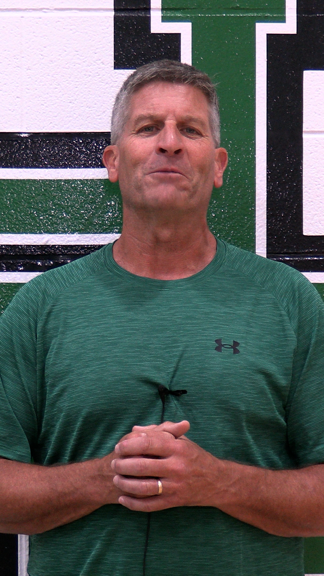 A man stands in front of a wall with green and black stripes. He wears a green shirt with a logo.
