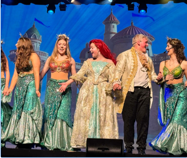 Six people on stage in costumes. Four women in green dresses with two men, one wearing a gold costume.