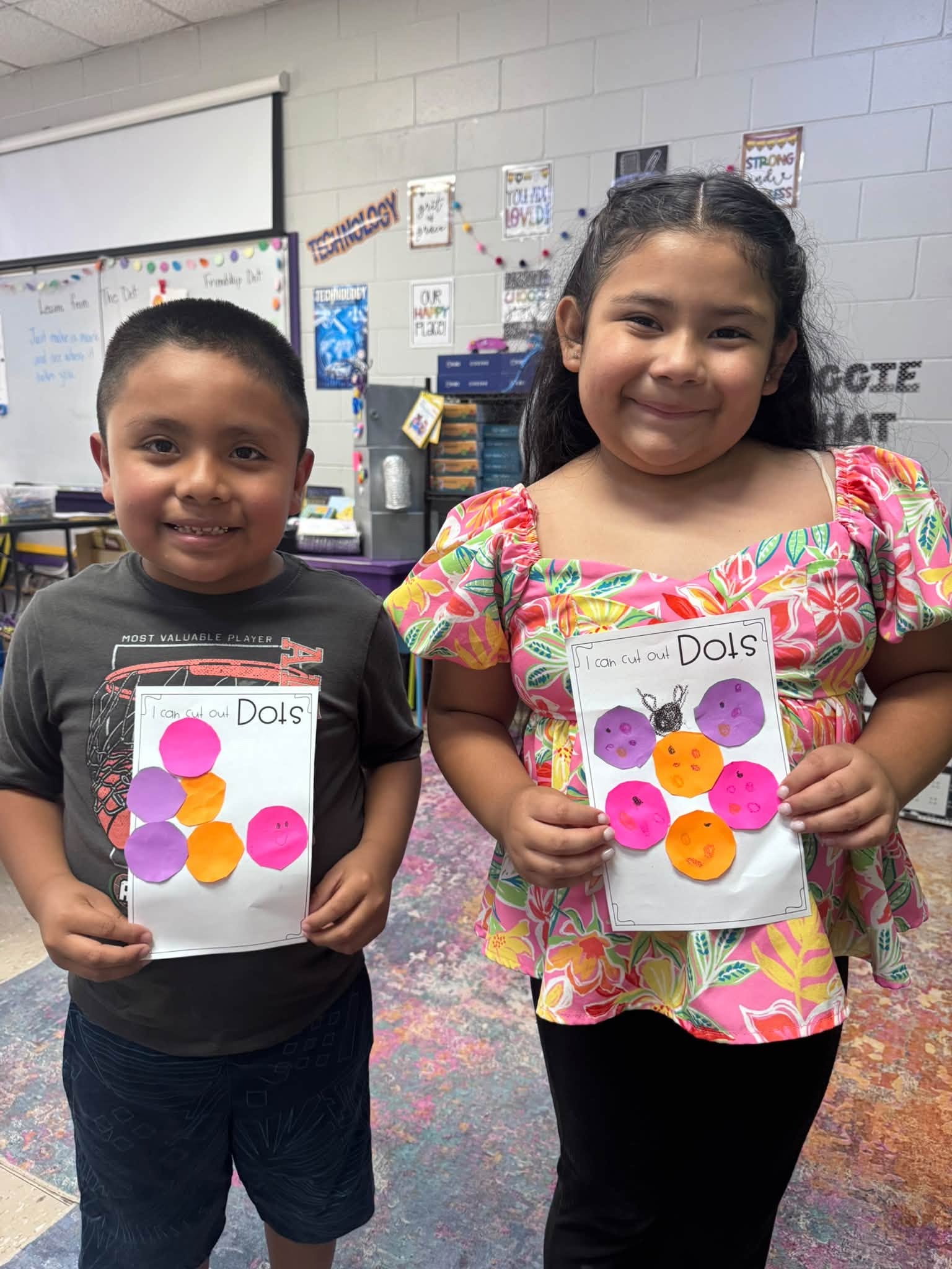 Two children stand in a classroom holding papers with circular images. A girl smiles, holding a paper with colorful circles. A boy beside her looks happy, holding similar papers.