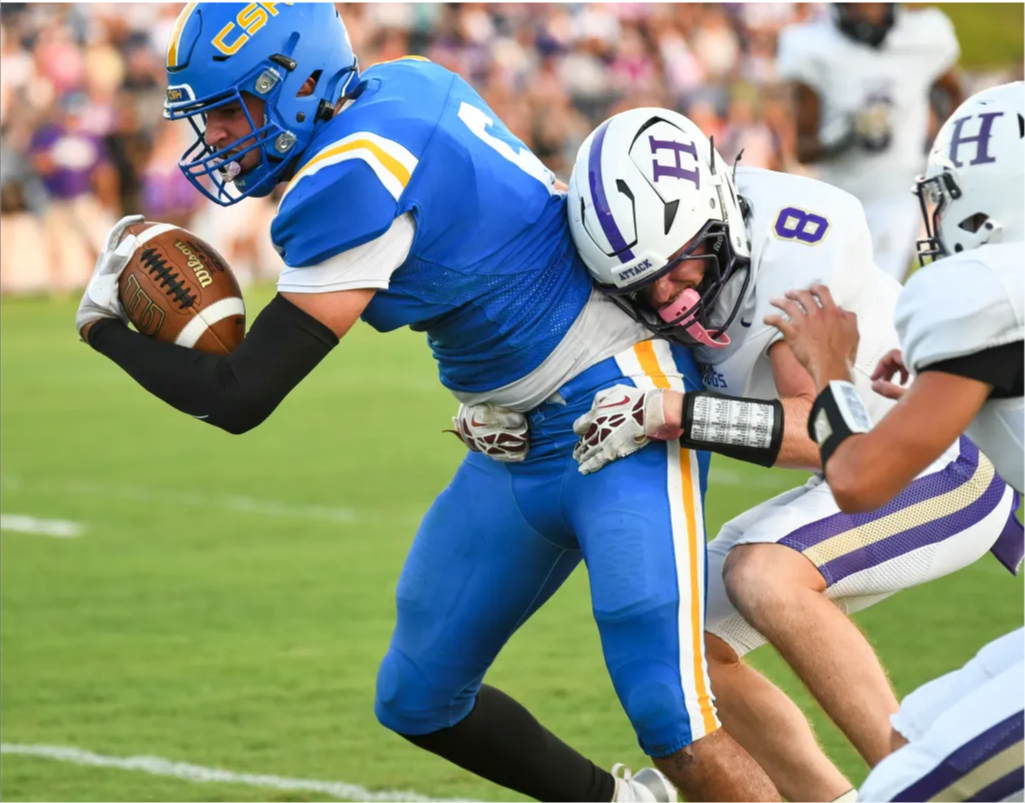 A football player in blue and white uniform runs with the ball as another player tackles him.