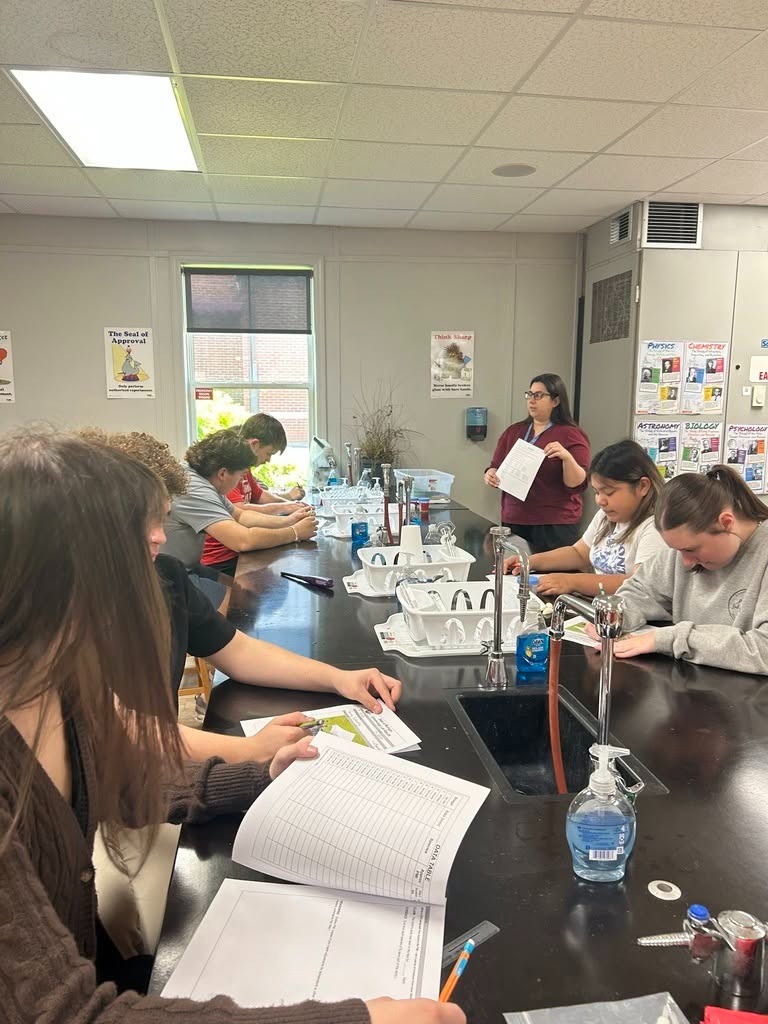 Lab room with students seated at tables, one holding papers and another holding a paper. A woman stands near a sink.
