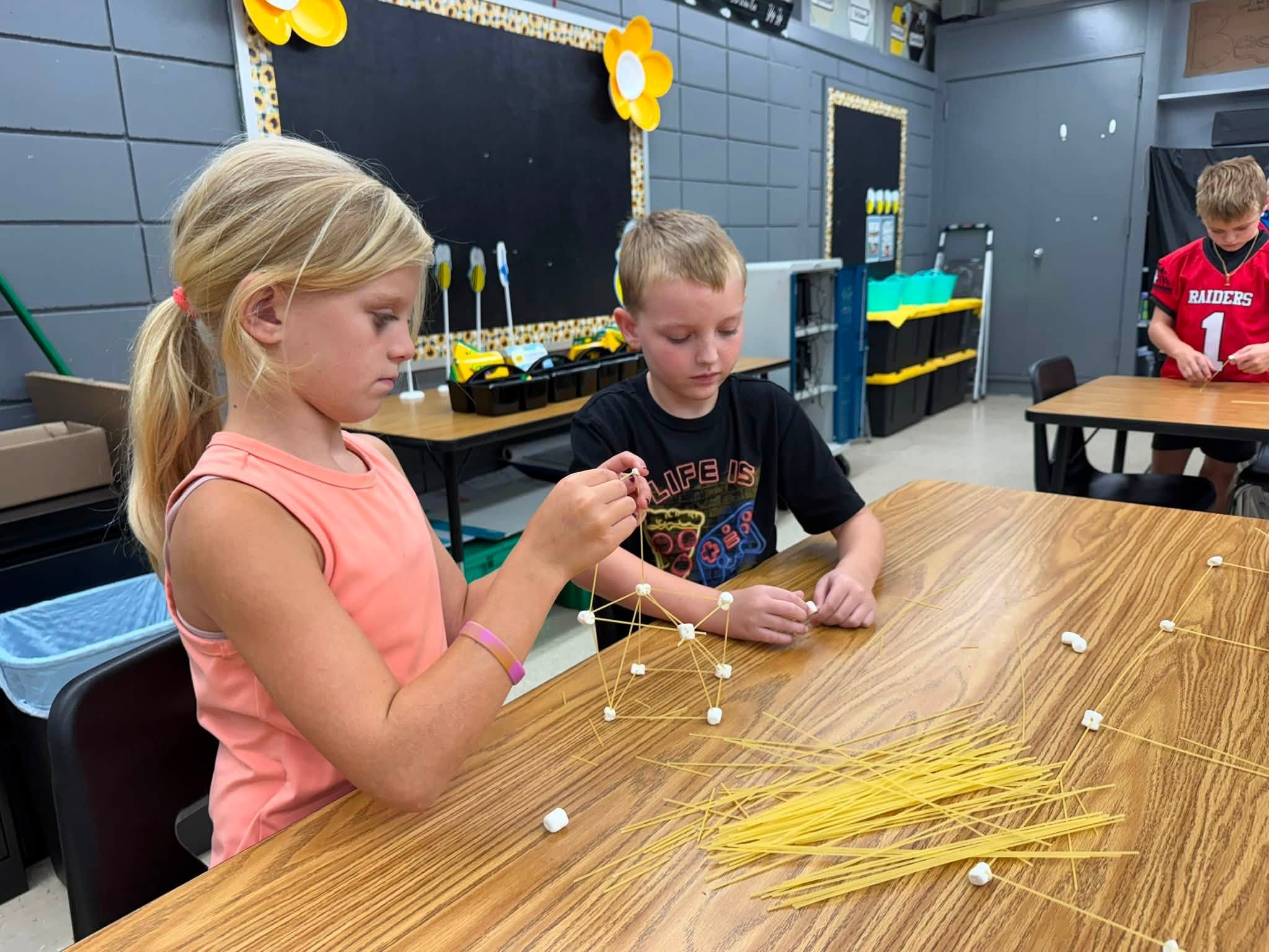 Two children, one with blonde hair and one with brown, sit at a table. They are making a structure with long yellow straws.
