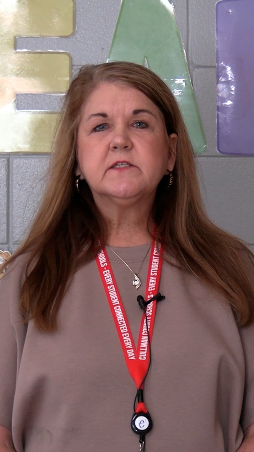 A woman with long hair and a red lanyard with white text stands in front of a colorful wall.
