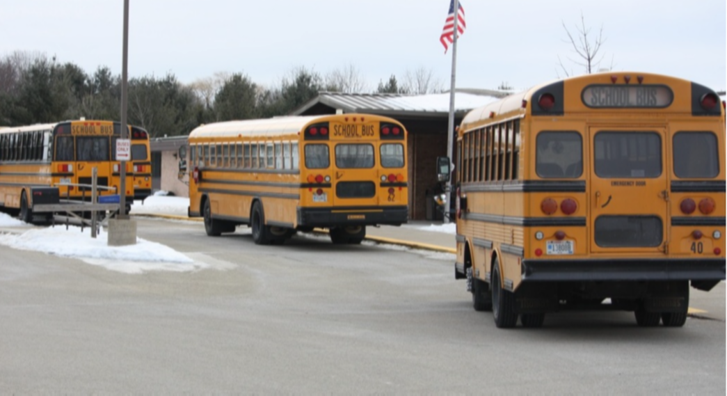 A line of school buses picture from the back. The buses are in front of a school and there is a flag flying from a flag pole in front of the school.