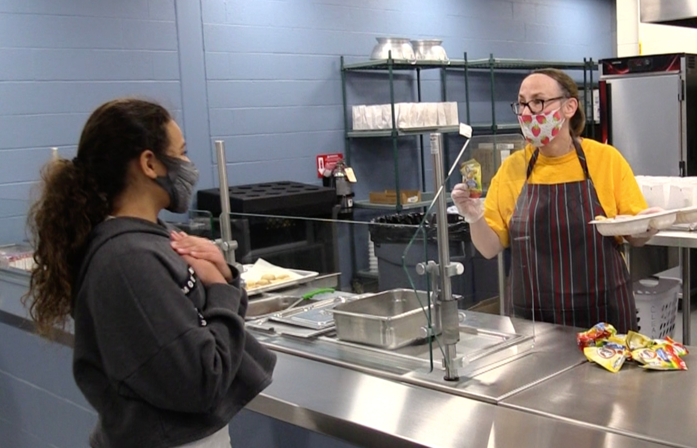 A student is getting food from a nutrition services employee. The student is holding her hands over her heart to thank the  employee for serving the meal to her.
