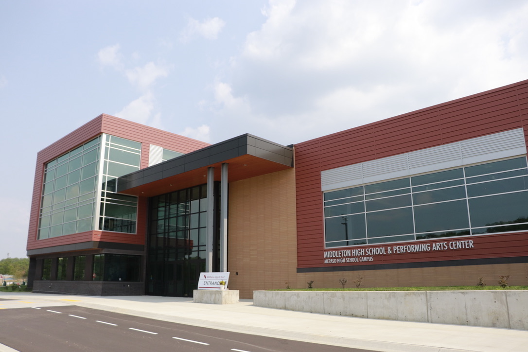 This is a picture showing the main entrance of the Middleton High School & Performing Arts Center. The building has a combination of materials & colors on the exterior, including red panels, sandy colored stone & lots of glass.