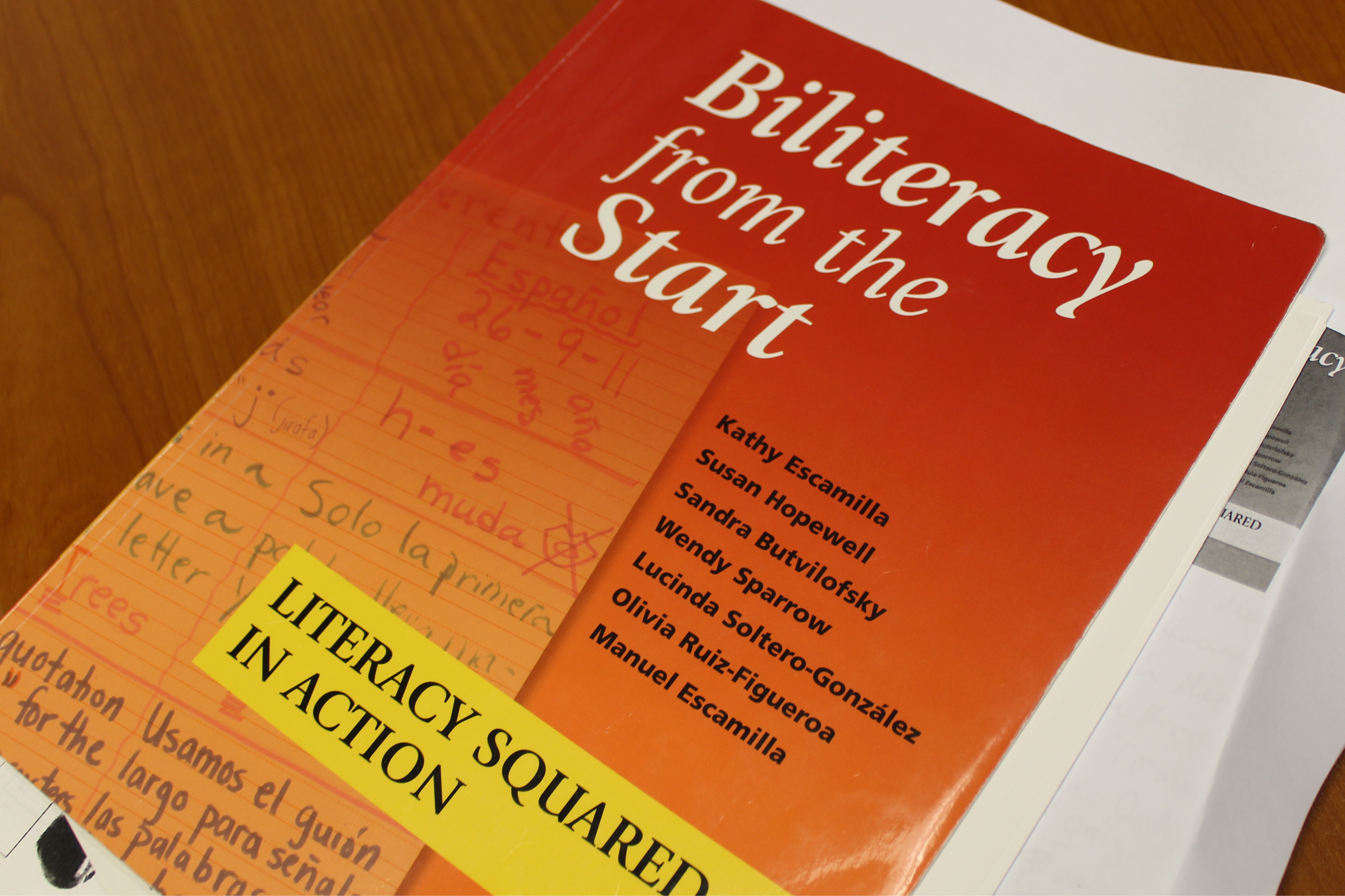 The image shows the front cover of a red book titled "Biliteracy from the Start" by Kathy Escamilla and several other authors. The cover design features a layered effect: The right side is a solid vibrant red with the title in a bold, white, serif font. The left side is a semi-transparent orange overlay showing handwritten classroom notes in Spanish and English on lined paper. A bright yellow rectangular sticker in the bottom left corner reads "LITERACY SQUARED IN ACTION" in bold black capital letters. The book is resting on a wooden surface alongside other white papers.