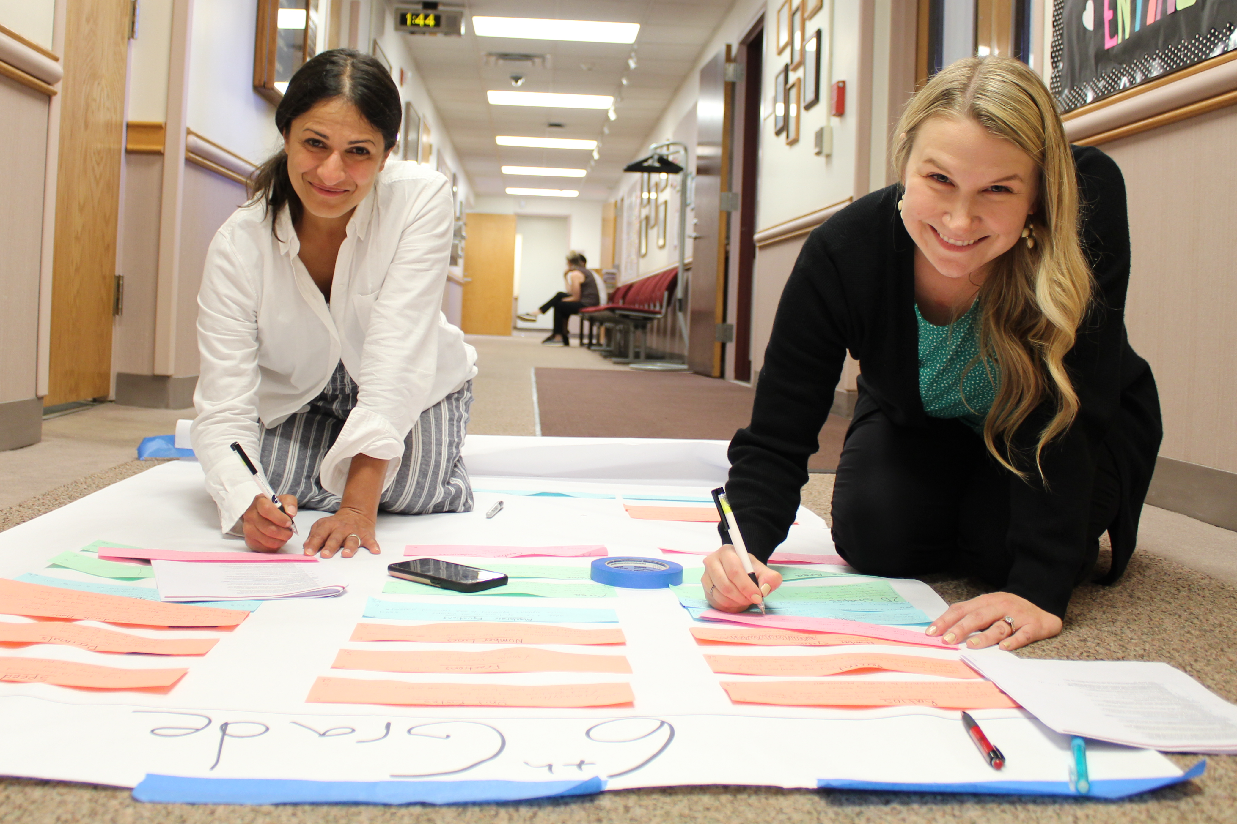 Two women kneel on a carpeted hallway floor, smiling as they collaborate on a large "6th Grade" curriculum mapping project spread out on white butcher paper. They are using markers to write on colorful pink, green, and blue paper strips taped to the poster, which include math-related headings like "Decimals" and "Fractions".