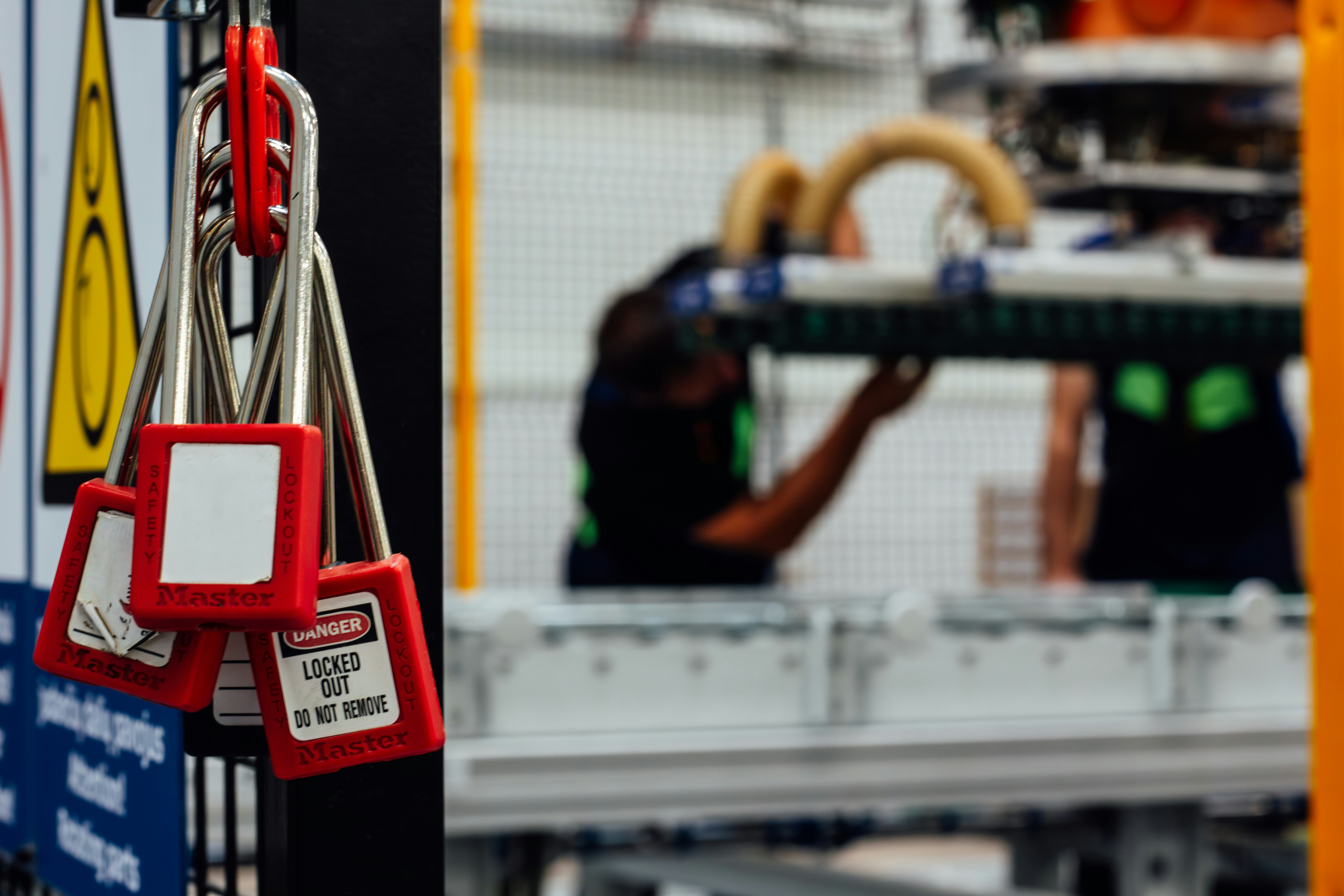 A close-up of several red Master Lock safety padlocks hanging on a black metal rack in an industrial setting. One lock clearly displays a white "DANGER: LOCKED OUT DO NOT REMOVE" label. In the background, workers in safety vests are blurred while performing maintenance on machinery.