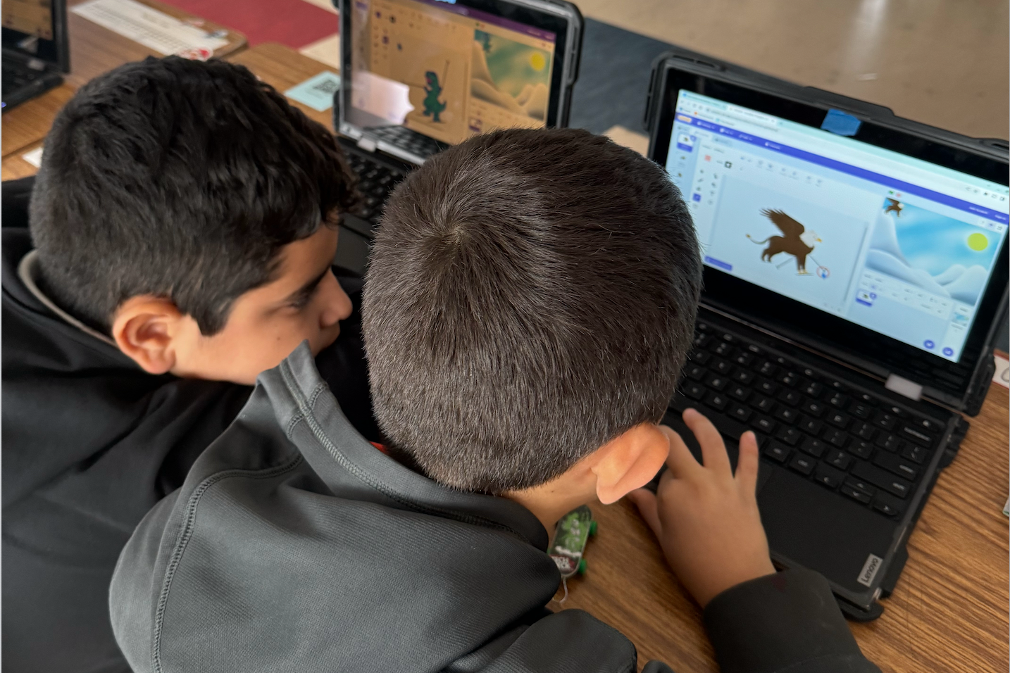 An over-the-shoulder view of two young students sitting side-by-side at a wooden desk, focused on a laptop screen. They are using a block-based coding program to animate a griffin character. A second laptop in the background shows a different student working on a similar project featuring a dinosaur character.