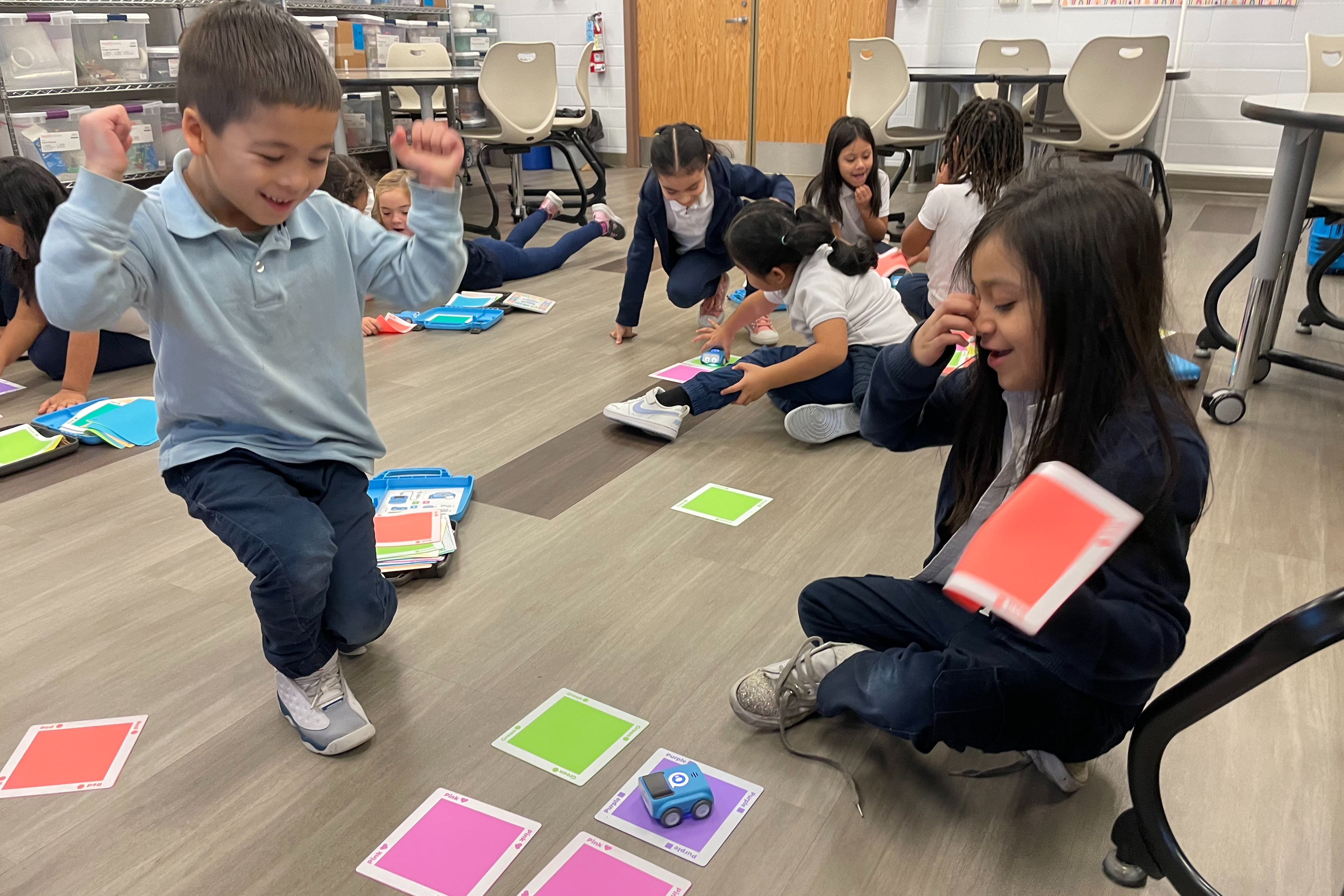 In a brightly lit classroom, a group of young students sits on the floor interacting with small, blue robotic cars. In the foreground, a boy in a light blue polo shirt cheers with his fists raised in excitement, while a girl sits nearby holding a red card. Other students are scattered in the background, focused on navigating their robots across colorful square floor tiles.