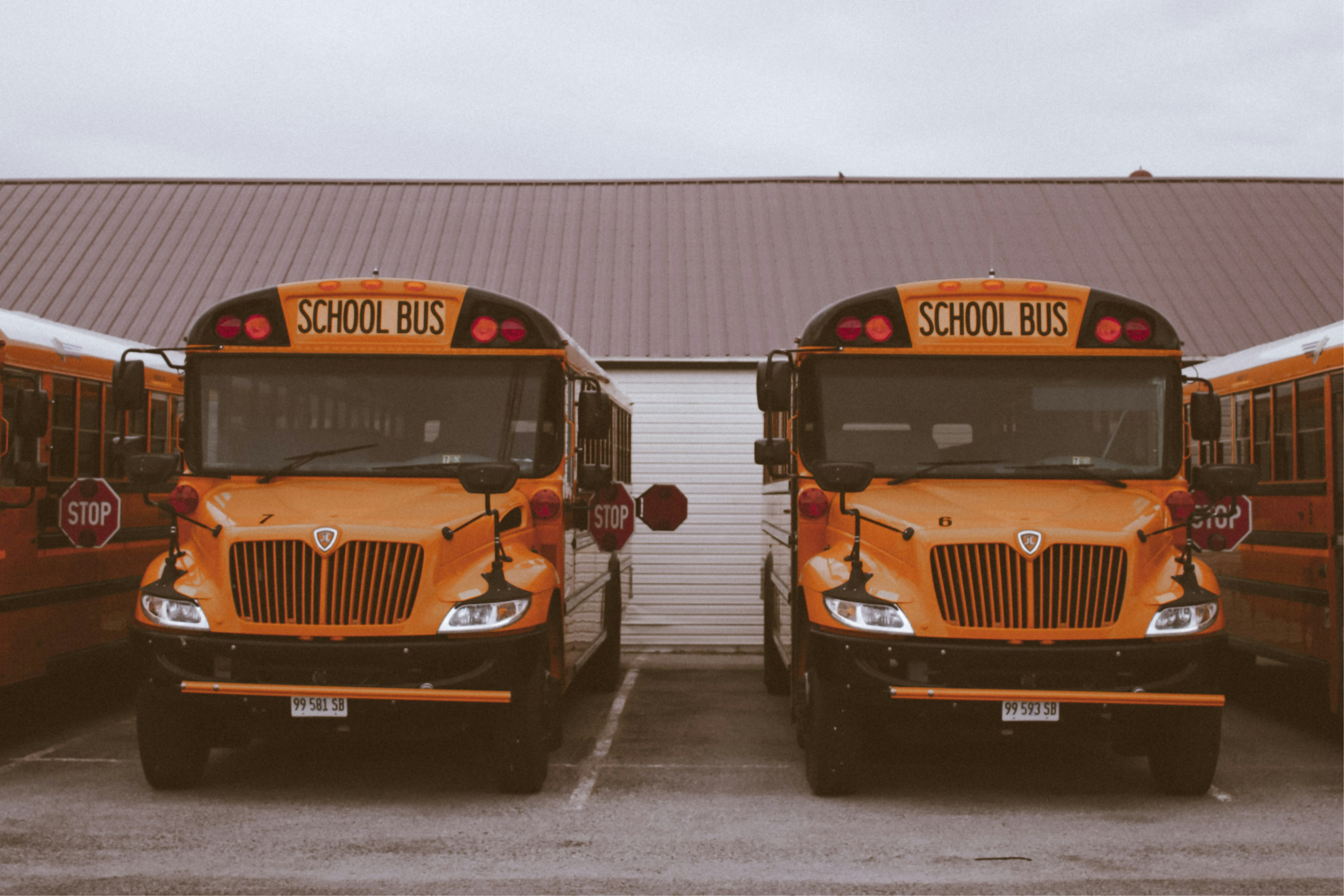 A symmetrical front-facing shot of two yellow school buses parked side-by-side in a lot under an overcast sky. The buses feature the "SCHOOL BUS" sign in bold black letters above the windshield and a vertical ribbed grille. Parts of two additional buses are visible on the far left and right edges, all positioned in front of a long building with a brown corrugated metal roof.