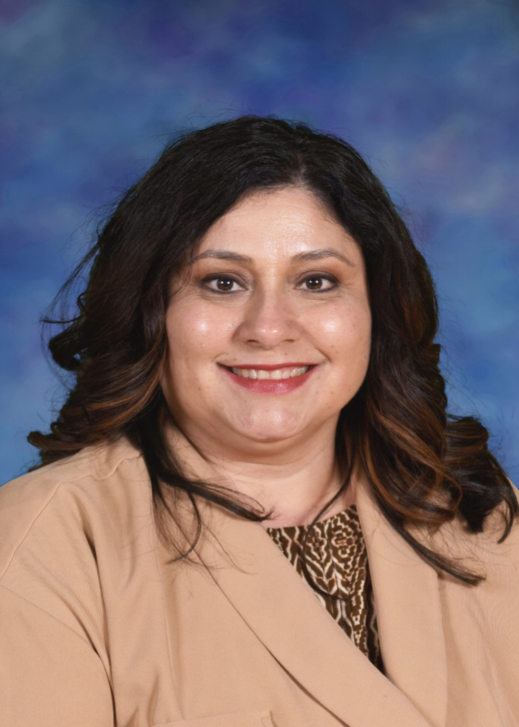 Headshot of a smiling woman with wavy dark brown hair, wearing a tan jacket over a patterned top against a blue background.