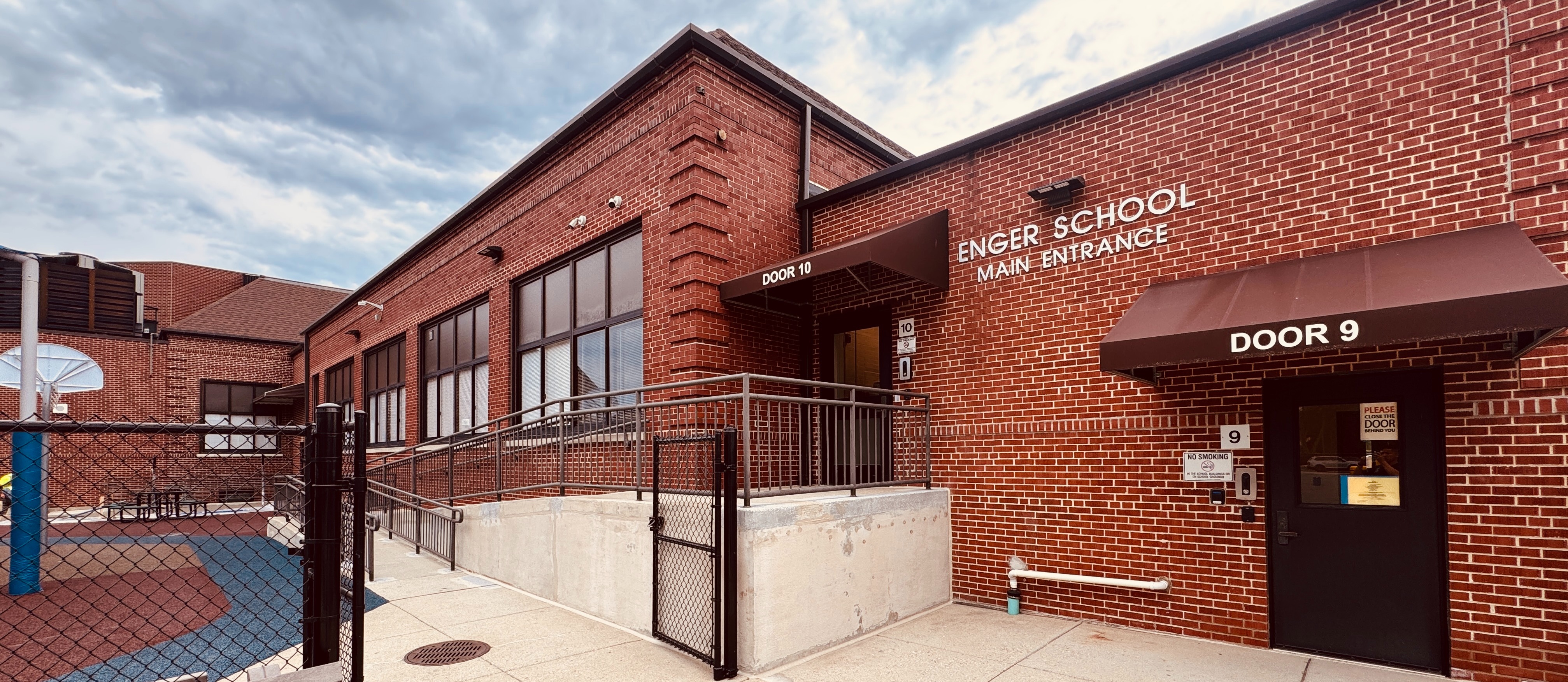 A wide-angle, exterior view of the Enger School Main Entrance shows a modern red brick building under a cloudy sky.  The entrance features two primary access points:  Door 9: A black door under a brown awning labeled "DOOR 9."  Door 10: Accessible via a concrete ramp with metal railings, located under a brown awning labeled "DOOR 10."  The "ENGER SCHOOL MAIN ENTRANCE" sign is clearly visible in white lettering on the brick wall between the two doors. To the left, a fenced-in playground area with blue and reddish-brown safety surfacing and a basketball hoop is visible. A wooden bench sits in the foreground, completing the clean and accessible school exterior.