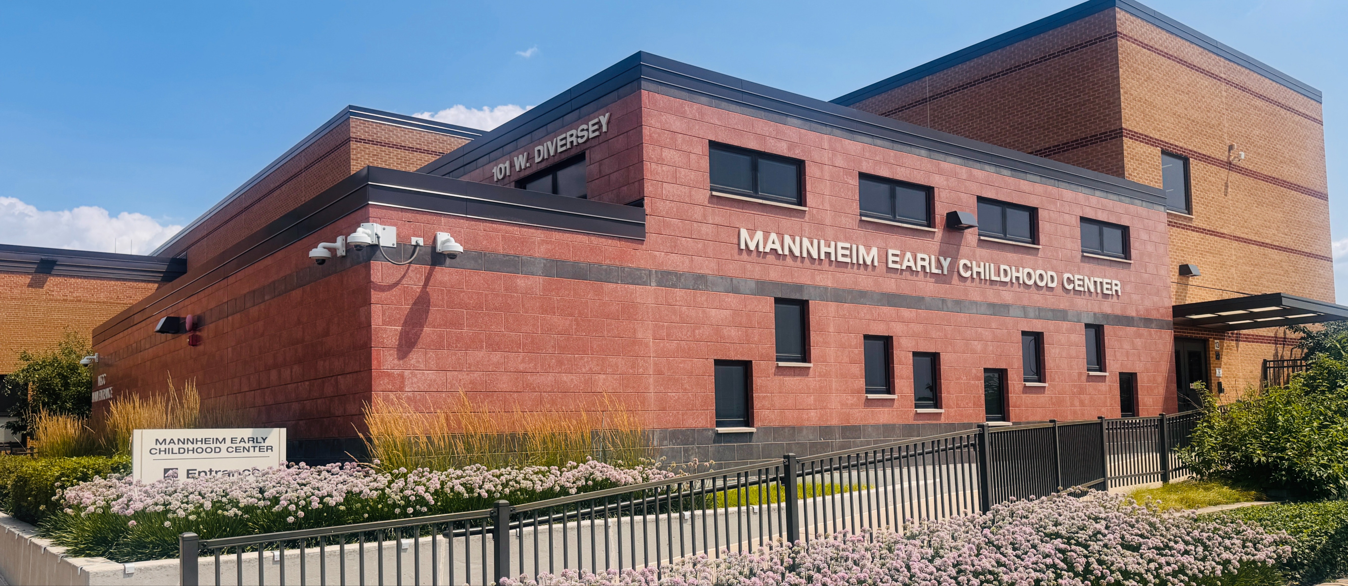 An exterior view of the Mannheim Early Childhood Center, a modern two-story building featuring a red masonry facade with dark grey accents. The building's address, 101 W. Diversey, is displayed in silver lettering above the main signage.  A black metal fence runs along a concrete walkway in the foreground, bordered by neatly landscaped beds of light purple flowers and tall ornamental grasses. The scene is set under a clear blue sky, highlighting the clean lines and contemporary architectural design of the facility.
