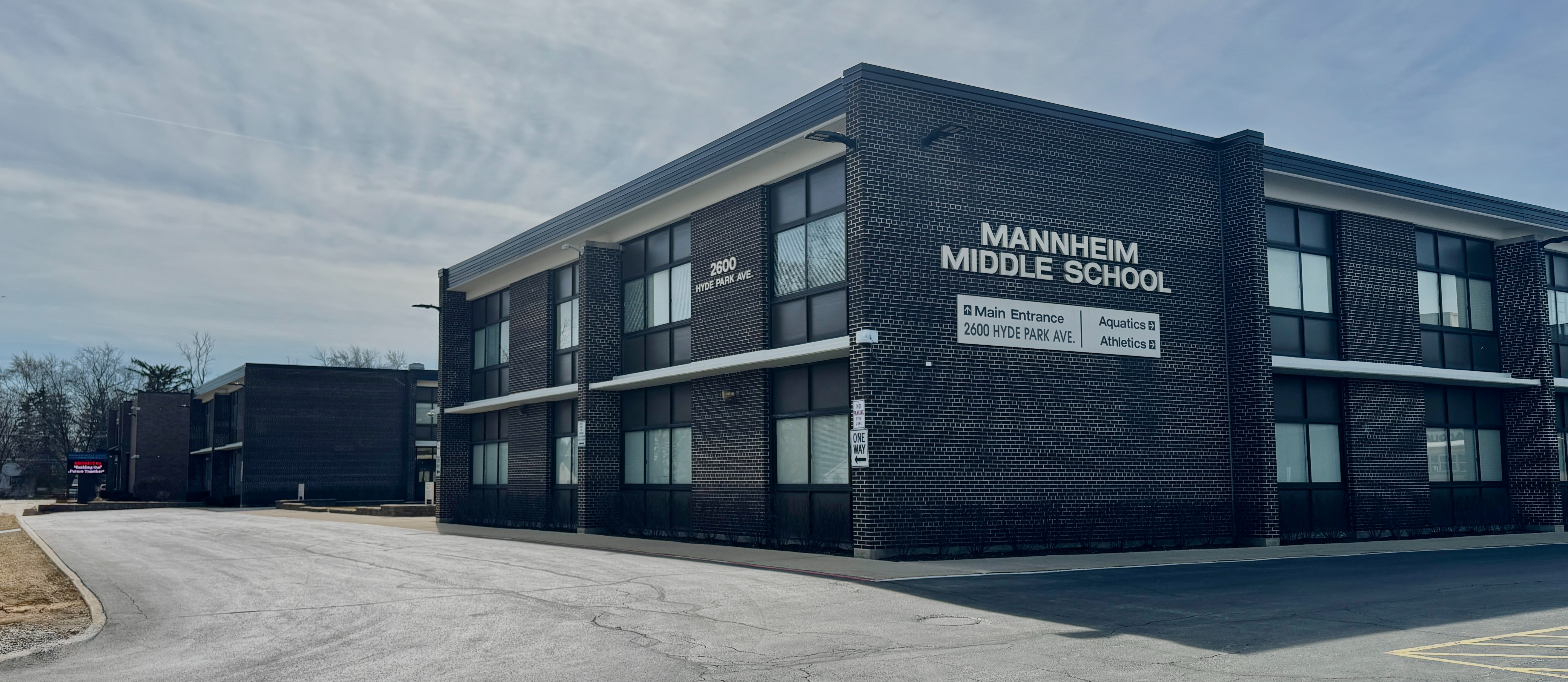 An exterior view of Mannheim Middle School, a large two-story building with a dark brick facade and expansive glass windows. The silver signage on the side of the building clearly displays the school name and its address, 2600 Hyde Park Ave, with directional indicators for the Main Entrance, Aquatics, and Athletics. The school is situated alongside a paved parking area and walkways under an overcast sky.