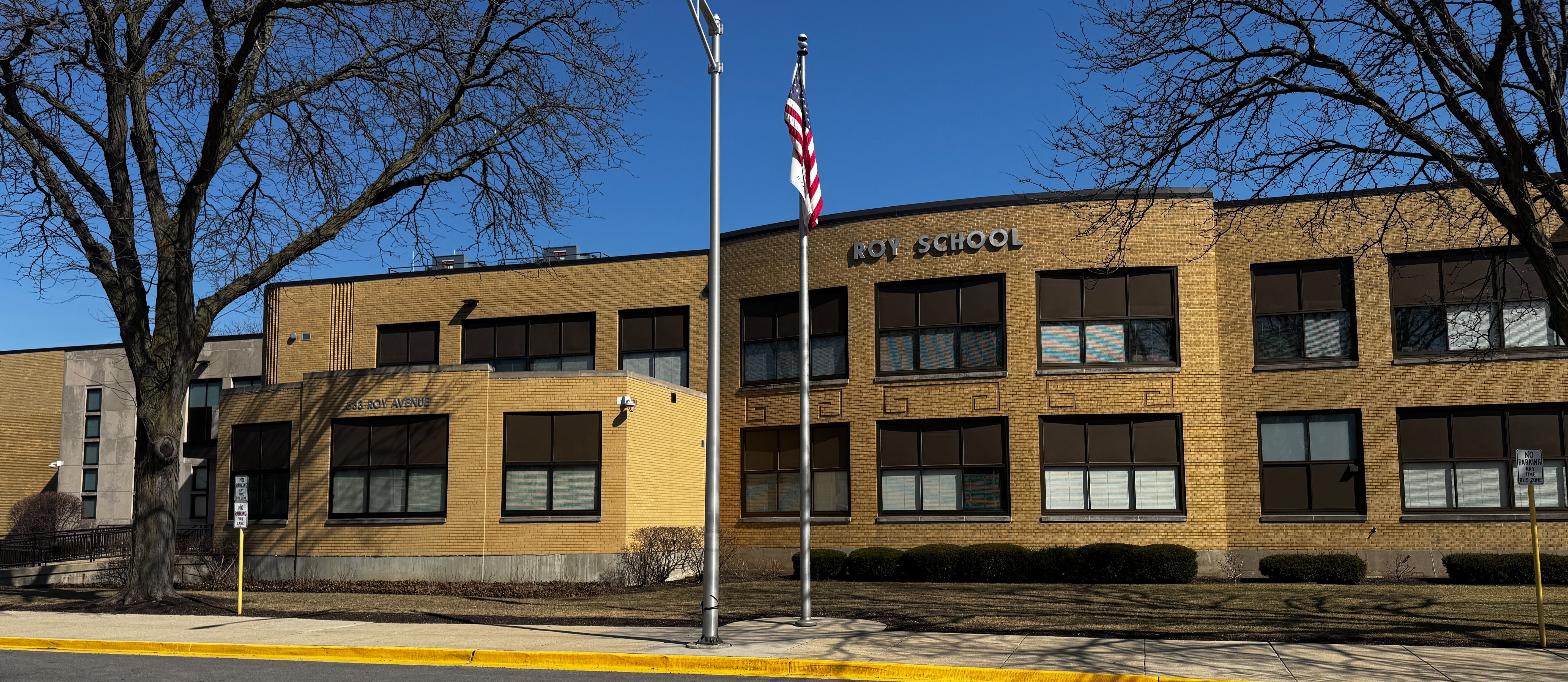 A wide, eye-level shot of the Roy School exterior, a two-story tan brick building with many large, dark-framed windows. An American flag flies from a tall silver flagpole in the center foreground, set against a clear blue sky. Bare winter trees frame the building on both sides, and a yellow-painted curb runs along the bottom of the frame next to the sidewalk.