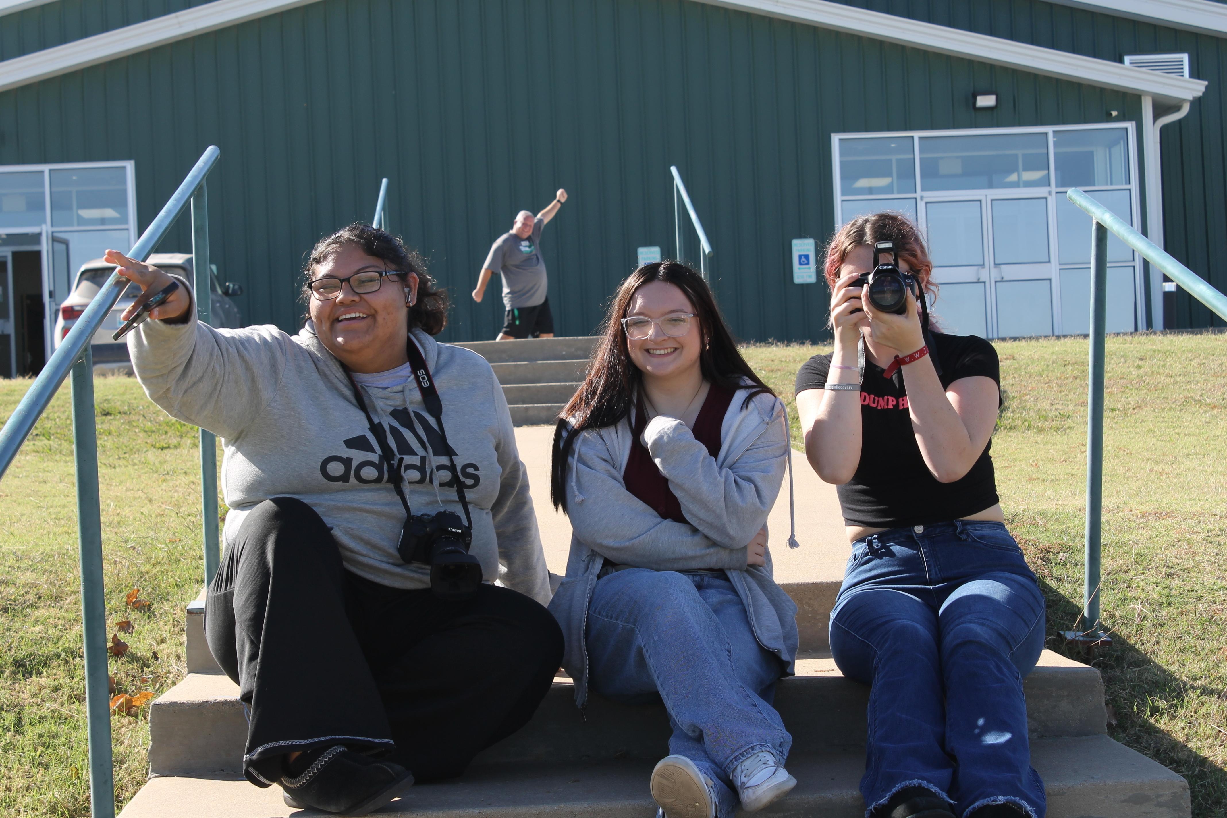 Journalism Staff cheesing while covering a softball game featuring our lovely principal Stacey Barton celebrating in the background. Pictured from left to right: Junior Mvhayv Salceda, Junior and Senior Editor Calora Cain, and Junior Maddyson Marson-Compton.