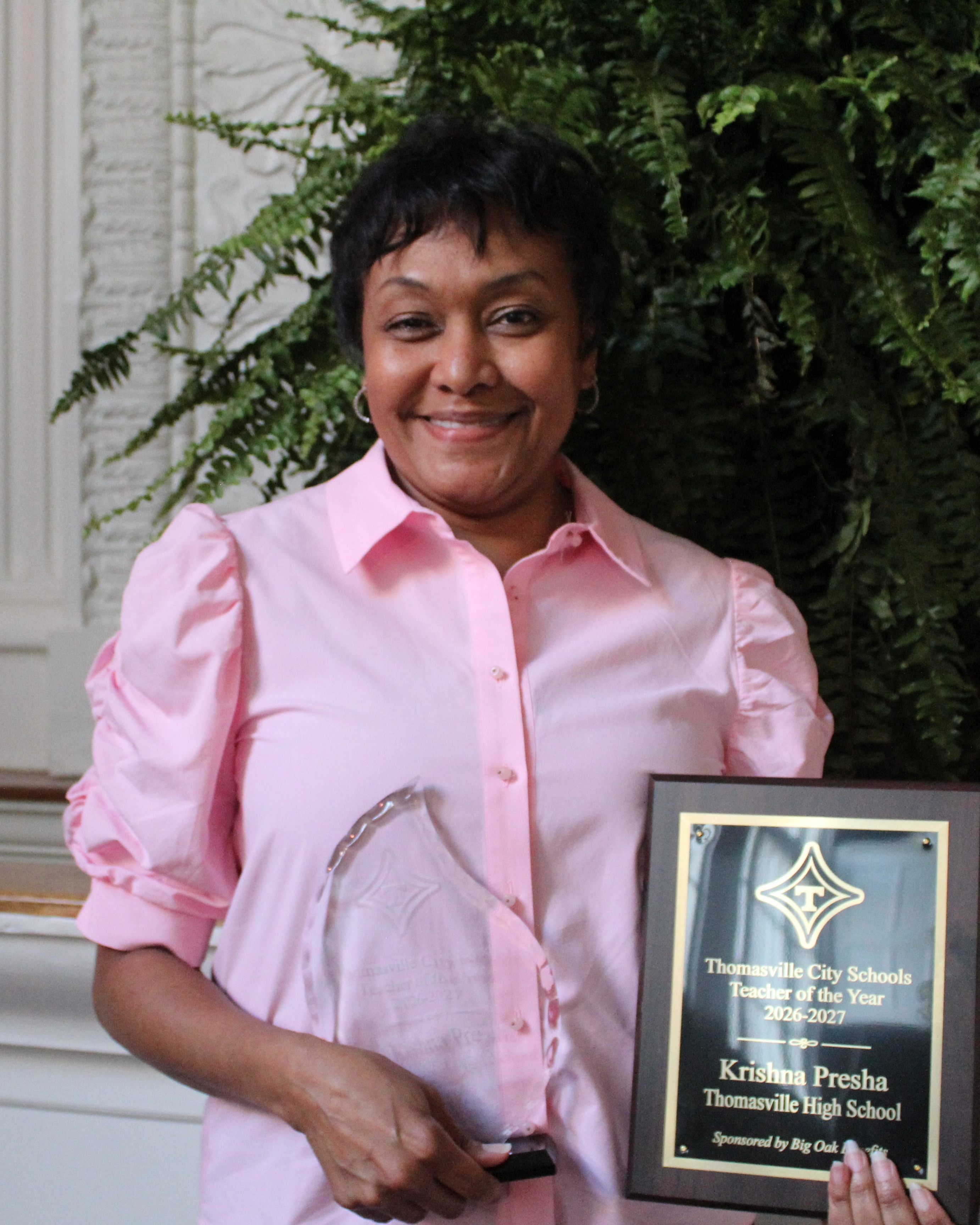 Woman in pink shirt holding a plaque and an award in front of a green plant.
